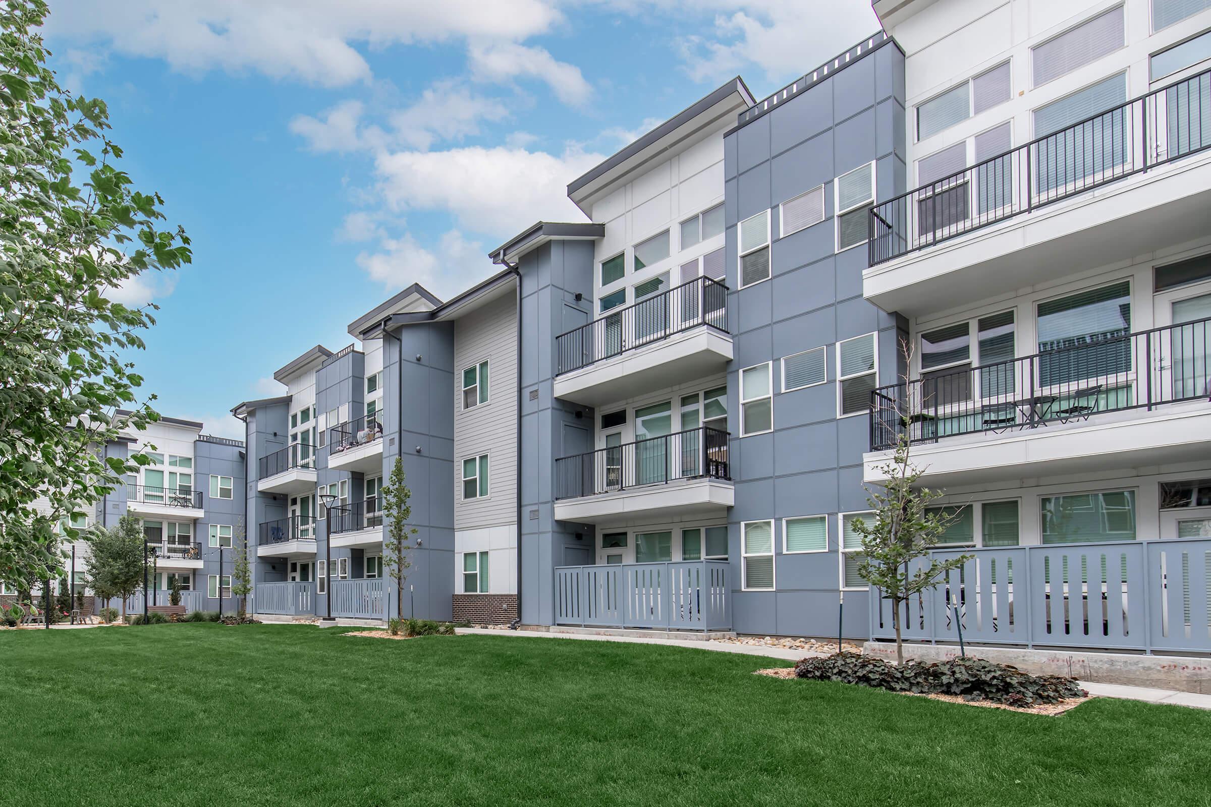 Modern apartment complex featuring three-story buildings with a sleek, gray exterior. The landscape includes manicured lawns, small trees, and decorative shrubs. Balconies are visible on upper levels, and large windows enhance the contemporary design. The sky is partly cloudy, adding a bright ambiance to the scene.