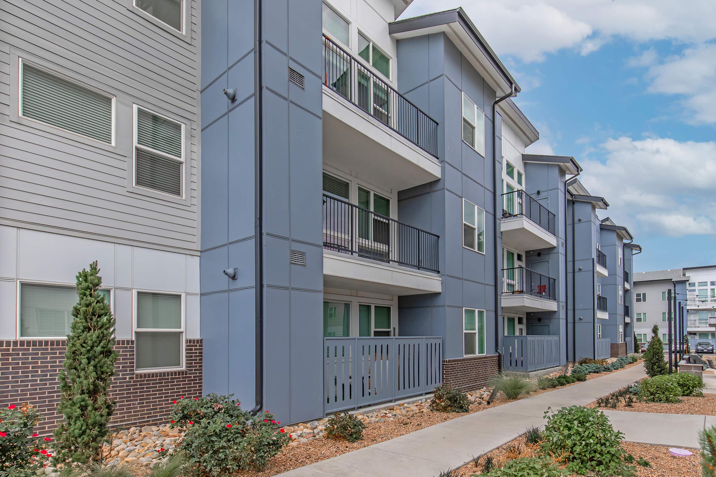A modern residential building featuring multiple apartments with balconies. The exterior is a mix of blue and gray colors, with brick accents and landscaped gardens along the walkway. Several windows are visible, and the sky is partly cloudy.