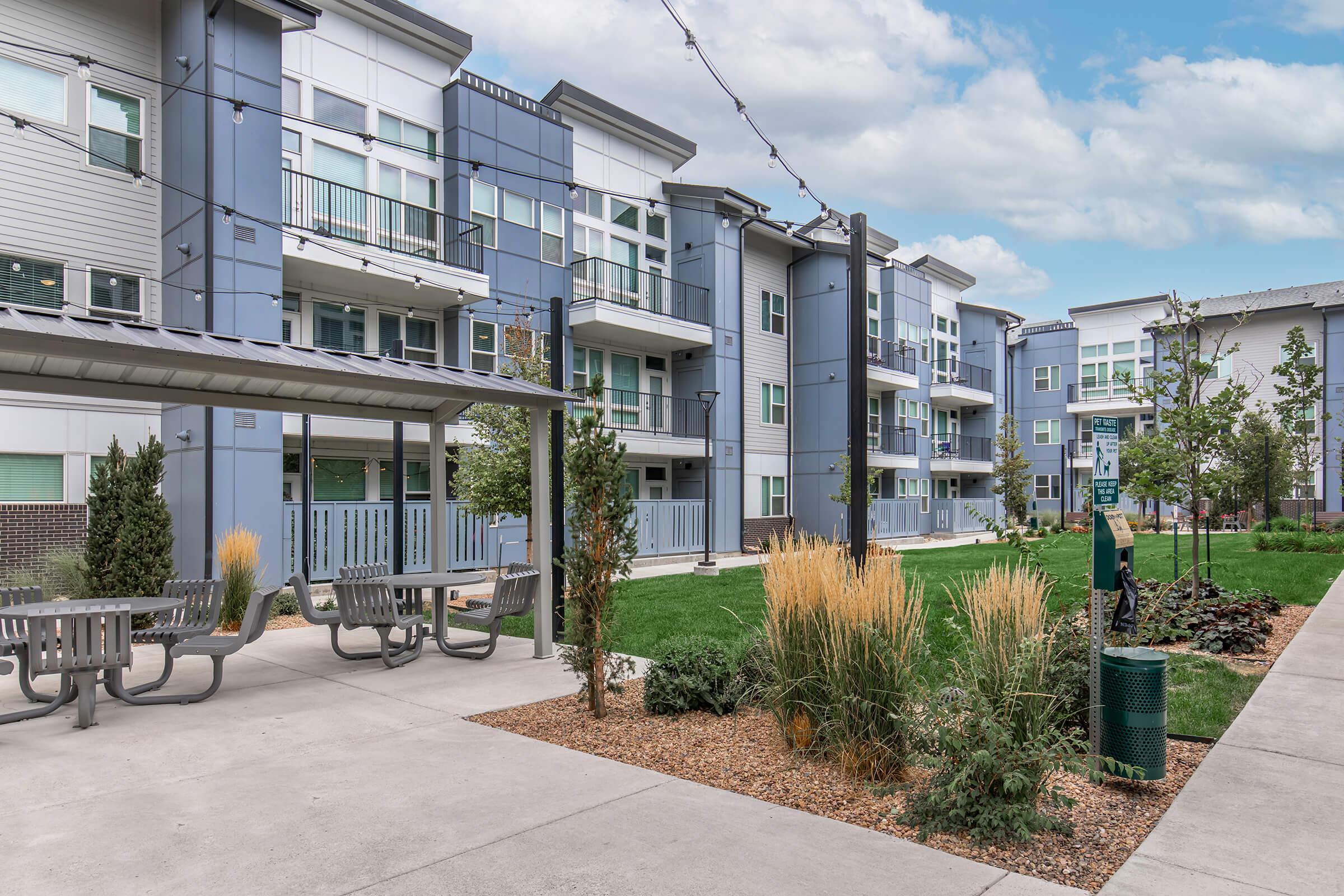 A modern residential complex featuring multiple gray apartment buildings. The area has landscaped green spaces with grasses and shrubs, along with seating areas and outdoor lighting. Pathways connect different sections, creating a welcoming community atmosphere.
