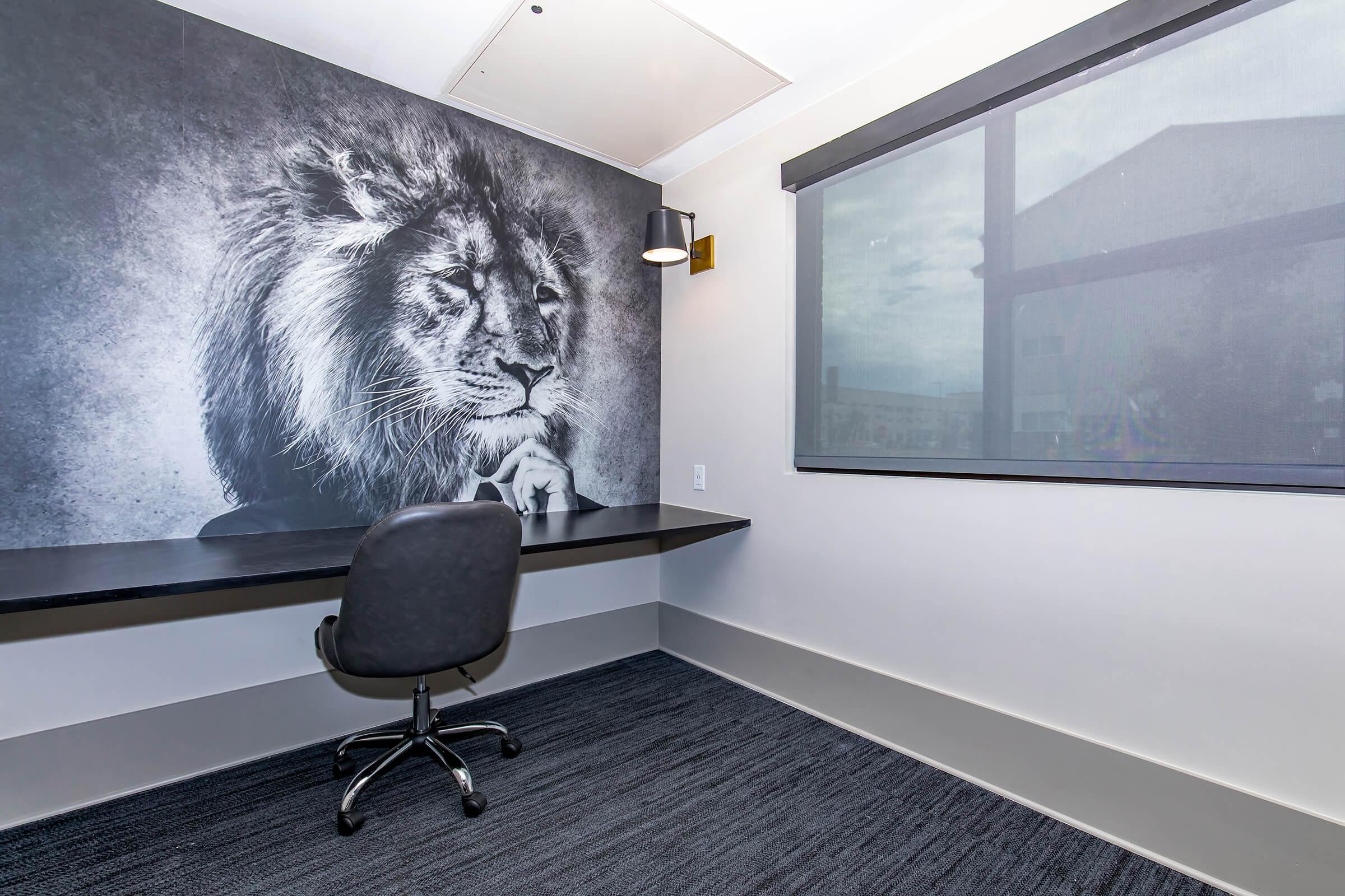 A minimalist workspace featuring a large black-and-white lion mural on one wall. A simple desk and a swivel chair are positioned in front of the mural, and a large window with a dark blind allows natural light into the room. The decor is modern and sleek, creating a calm atmosphere.
