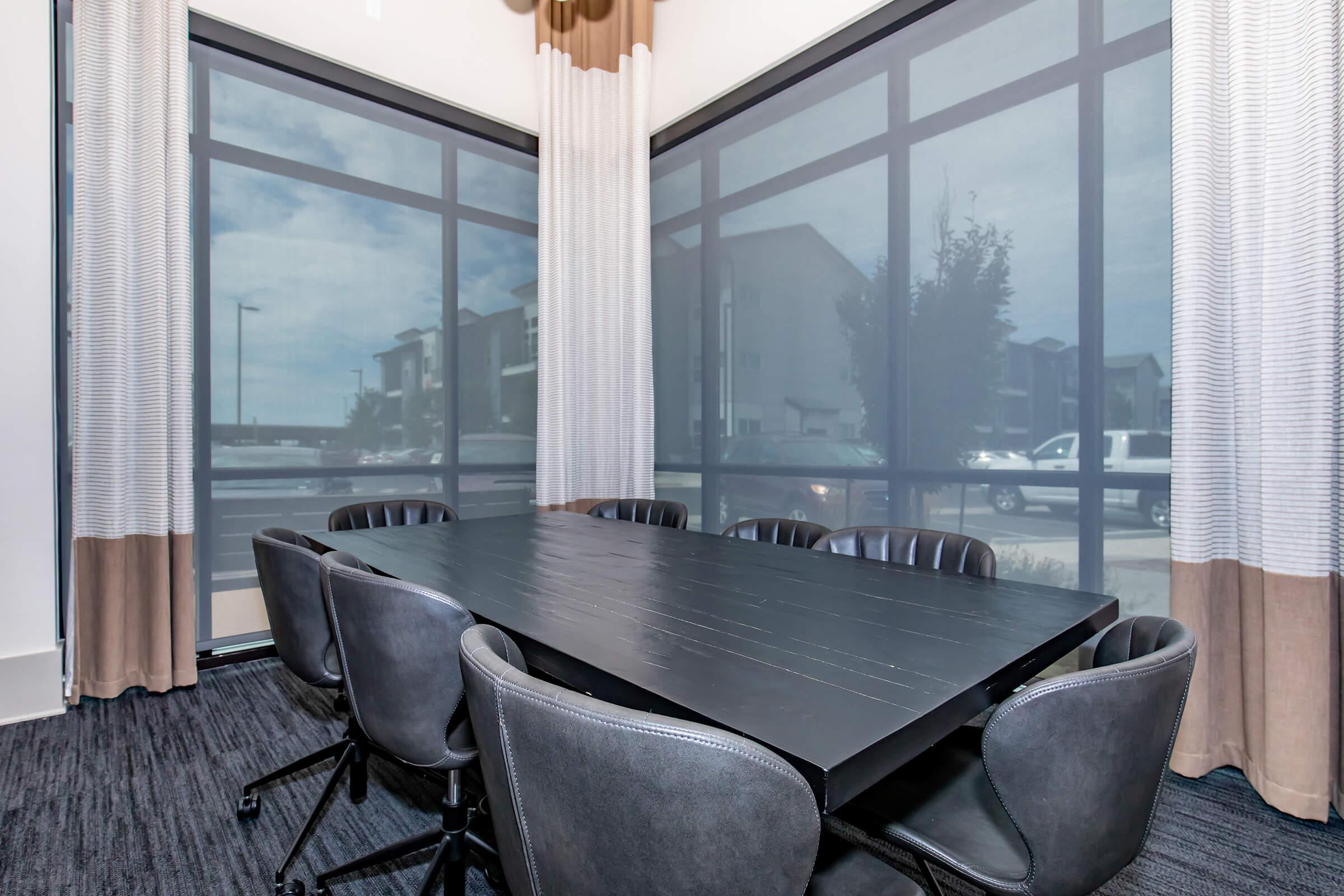 A modern conference room featuring a large black table surrounded by black rolling chairs. Large windows with sheer curtains provide natural light, reflecting a view of the outdoors. The room has a minimalist design with a dark carpet and a contemporary aesthetic.