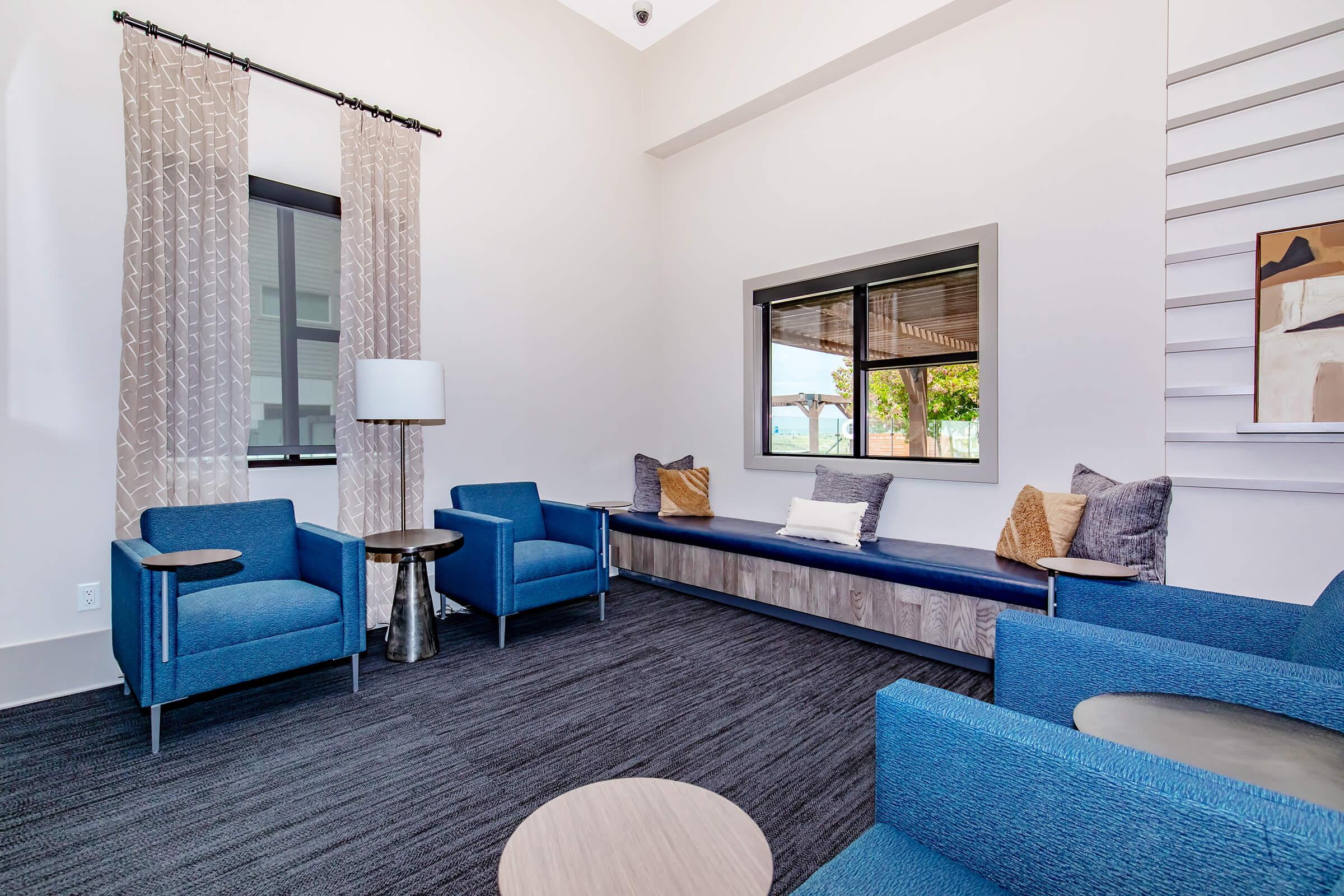 A modern seating area featuring three blue armchairs around a light wood circular table. There is a long, padded bench with decorative pillows against the wall, and a lamp on a side table. Large windows allow natural light to fill the space, complemented by neutral-colored curtains and artwork on the wall.