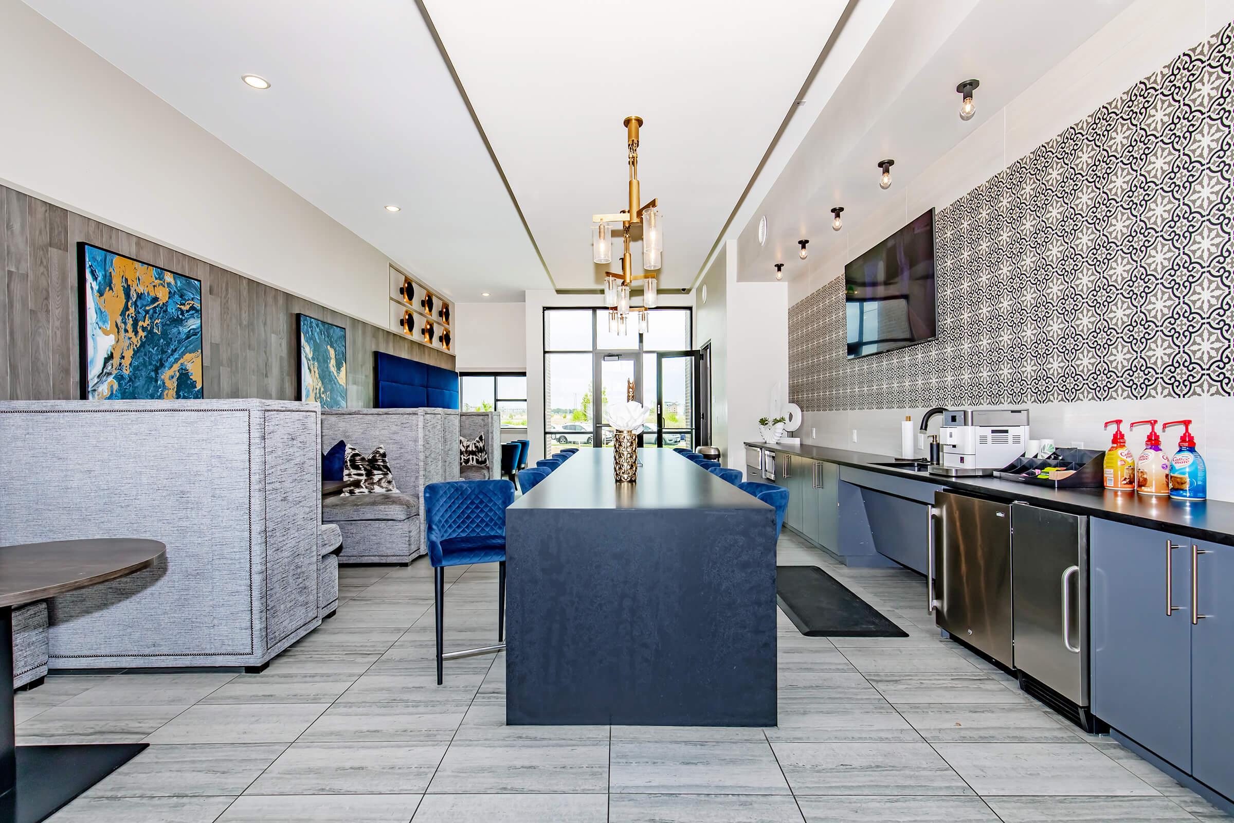 Modern interior of a stylish lounge area featuring a long, dark countertop with a coffee station, metal appliances, and decorative elements. Bright blue chairs are arranged around a table. The walls have a mix of patterned tiles and muted gray paneling, with ample natural light coming from large windows.