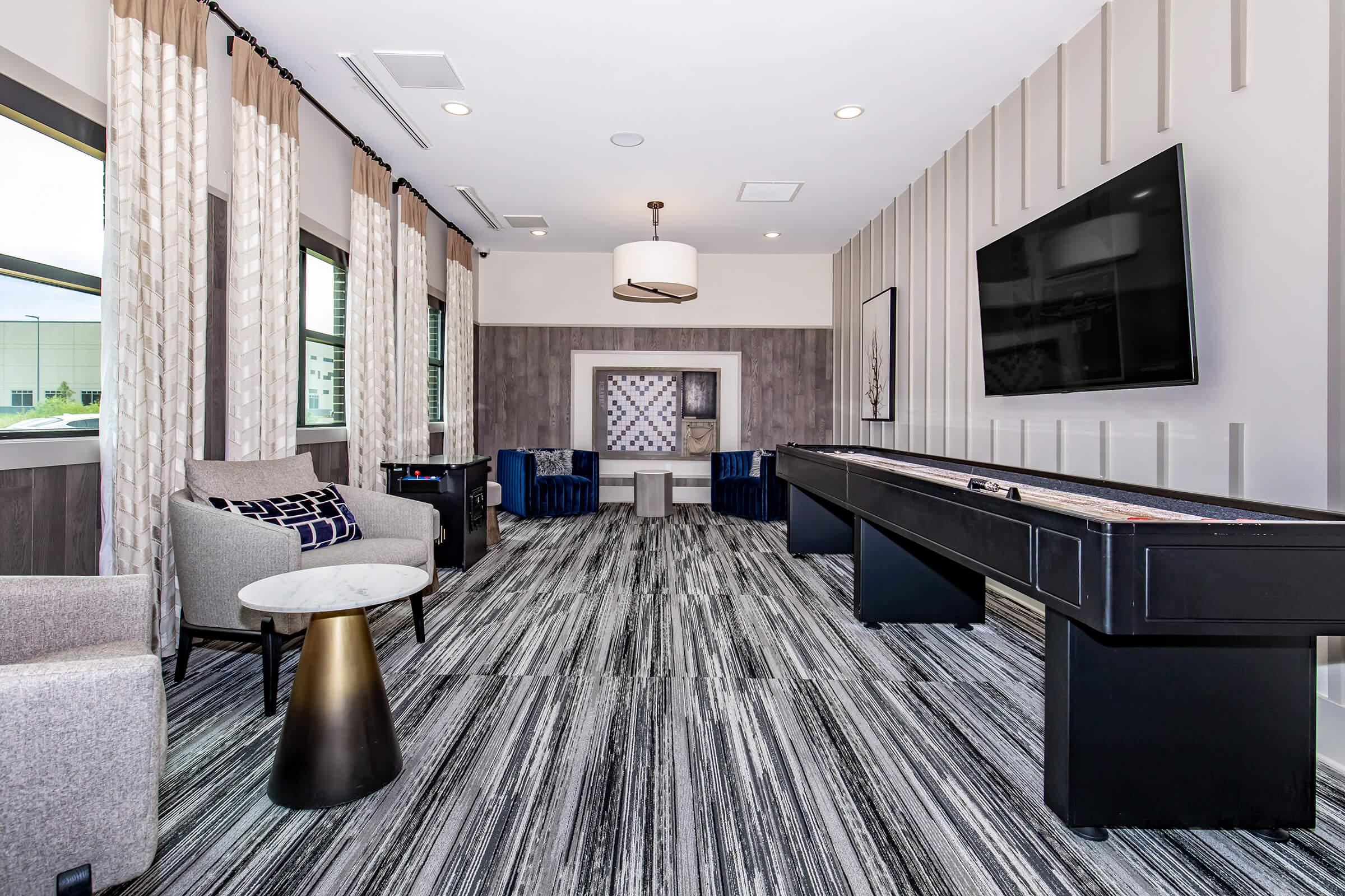 A modern lounge featuring two gray armchairs beside a small round table, a shuffleboard table in the center, and a large wall-mounted TV. Large windows are dressed with light curtains, and the floor has a striped gray pattern. The decor is sleek and contemporary, creating a welcoming atmosphere.