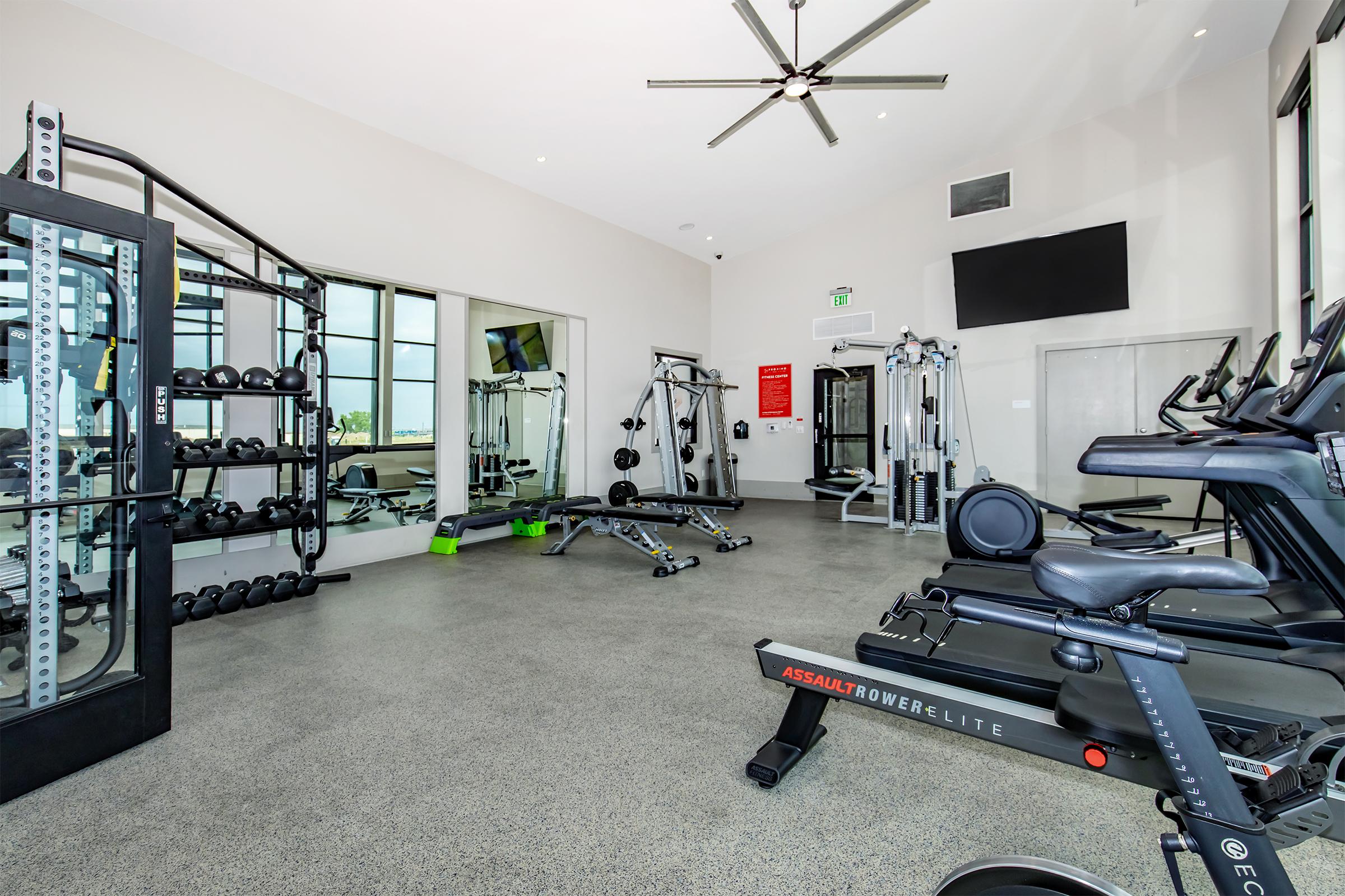 A modern gym interior featuring various workout equipment, including weight racks, exercise machines, and a rower. The space has large windows allowing natural light, a ceiling fan, and a wall-mounted TV. The floor is polished with a sleek finish, creating a clean and inviting atmosphere for fitness activities.