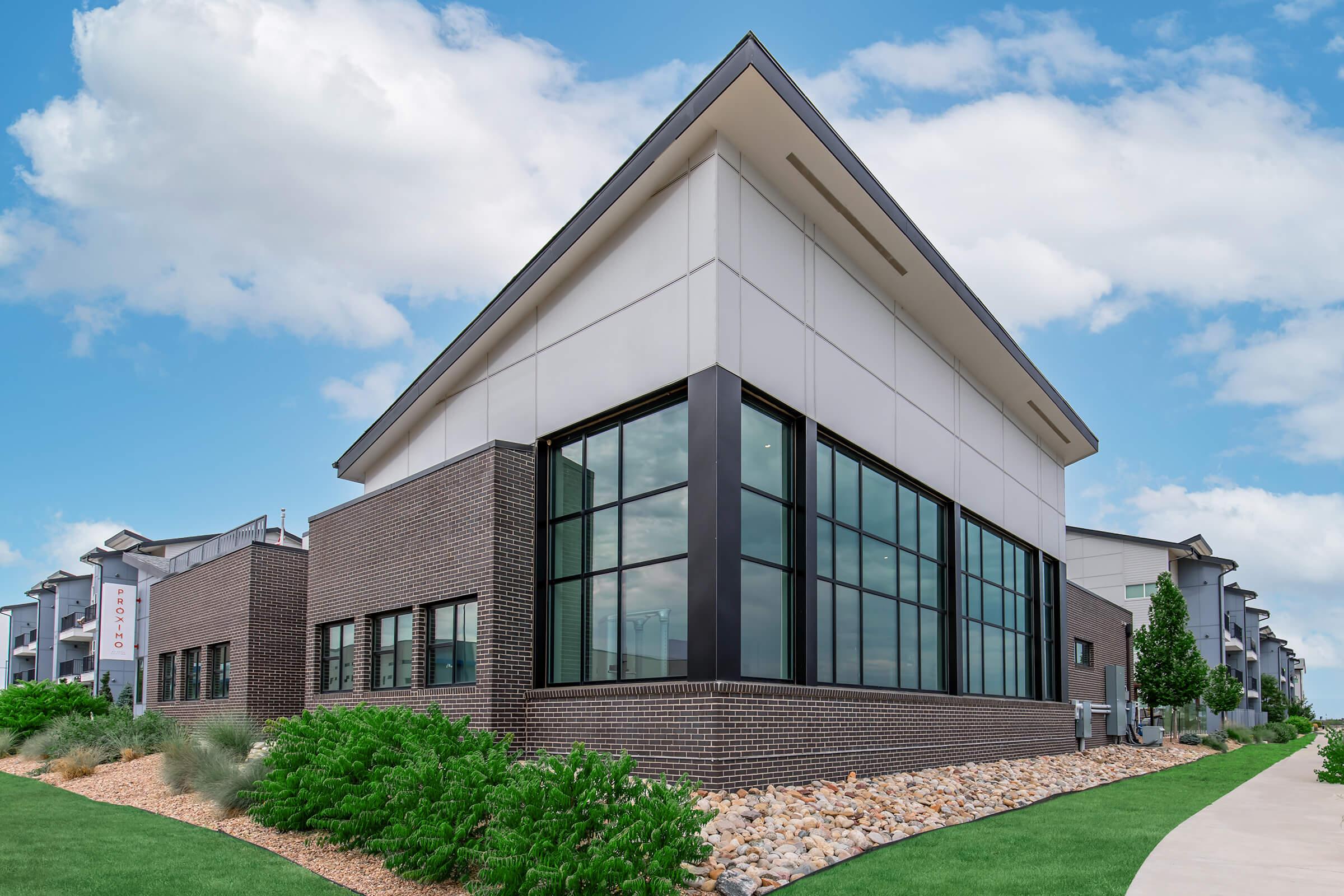 Modern building exterior featuring a mix of brick and light-colored paneling, large glass windows, and a sloped roof. The scene includes landscaped greenery and rocks in the foreground, under a partly cloudy blue sky. The architecture appears contemporary and stylish, typical of urban developments.