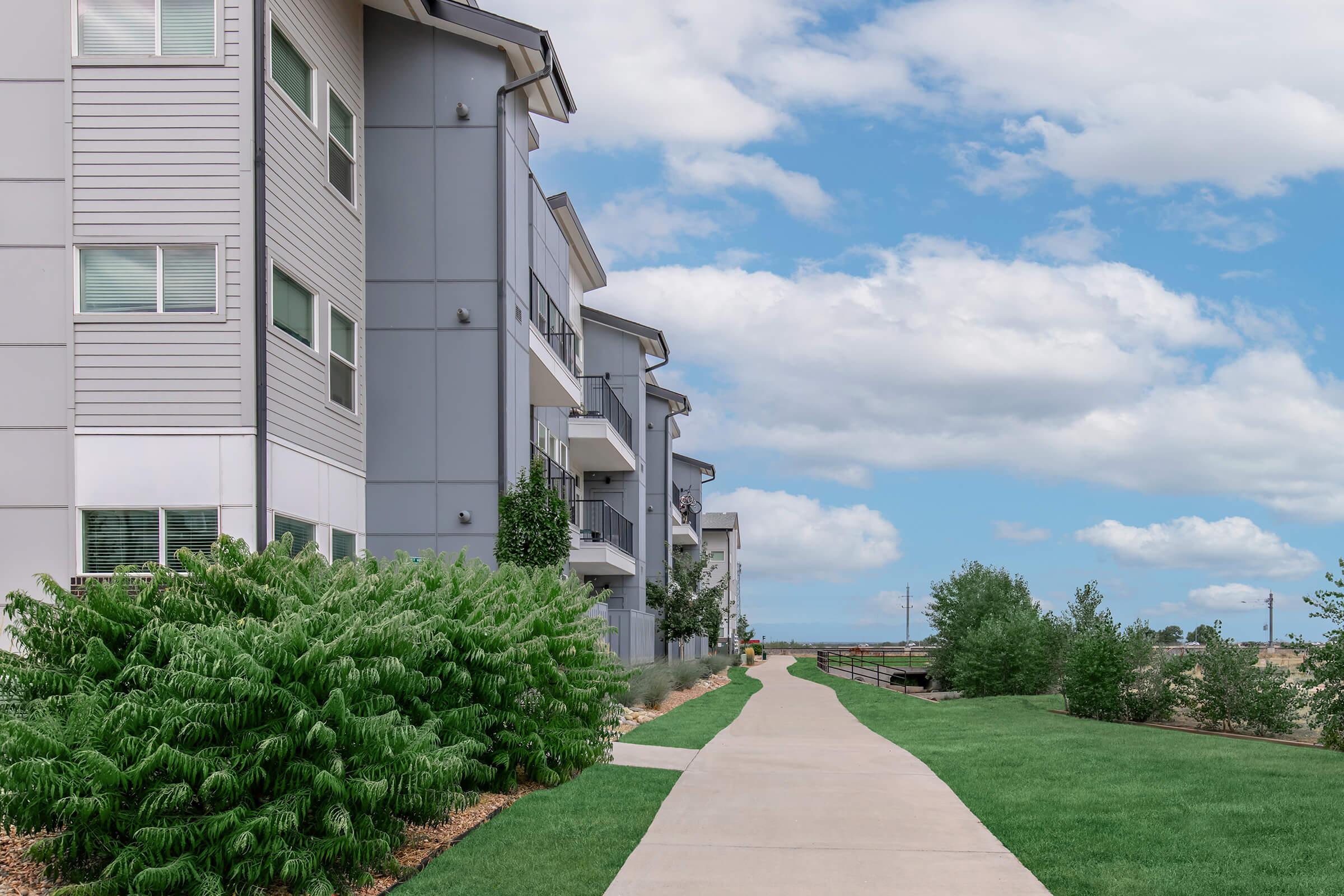 A landscaped walkway leads alongside modern apartment buildings, with green shrubs and grass on one side. The sky is partly cloudy, creating a pleasant atmosphere. The path appears well-maintained and inviting, bordered by nature and contemporary architecture.