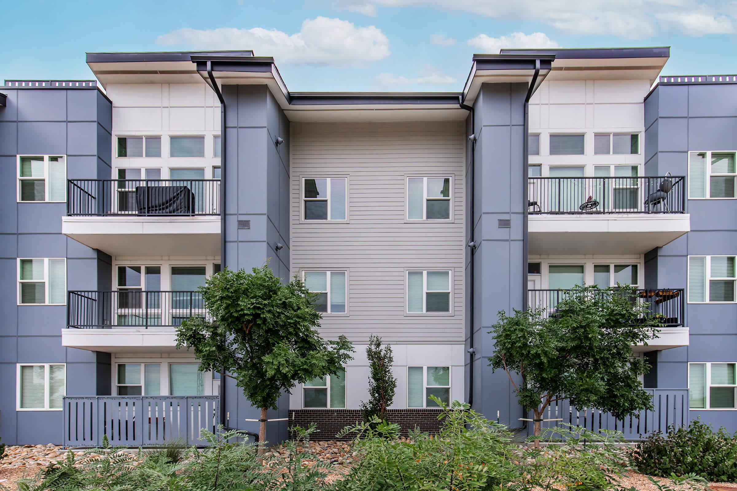 A modern apartment building with a symmetrical facade featuring two stories and multiple balconies. The exterior is primarily light grey with dark accents. There are trees and shrubs in the foreground, contributing to a landscaped environment. The sky is partly cloudy.