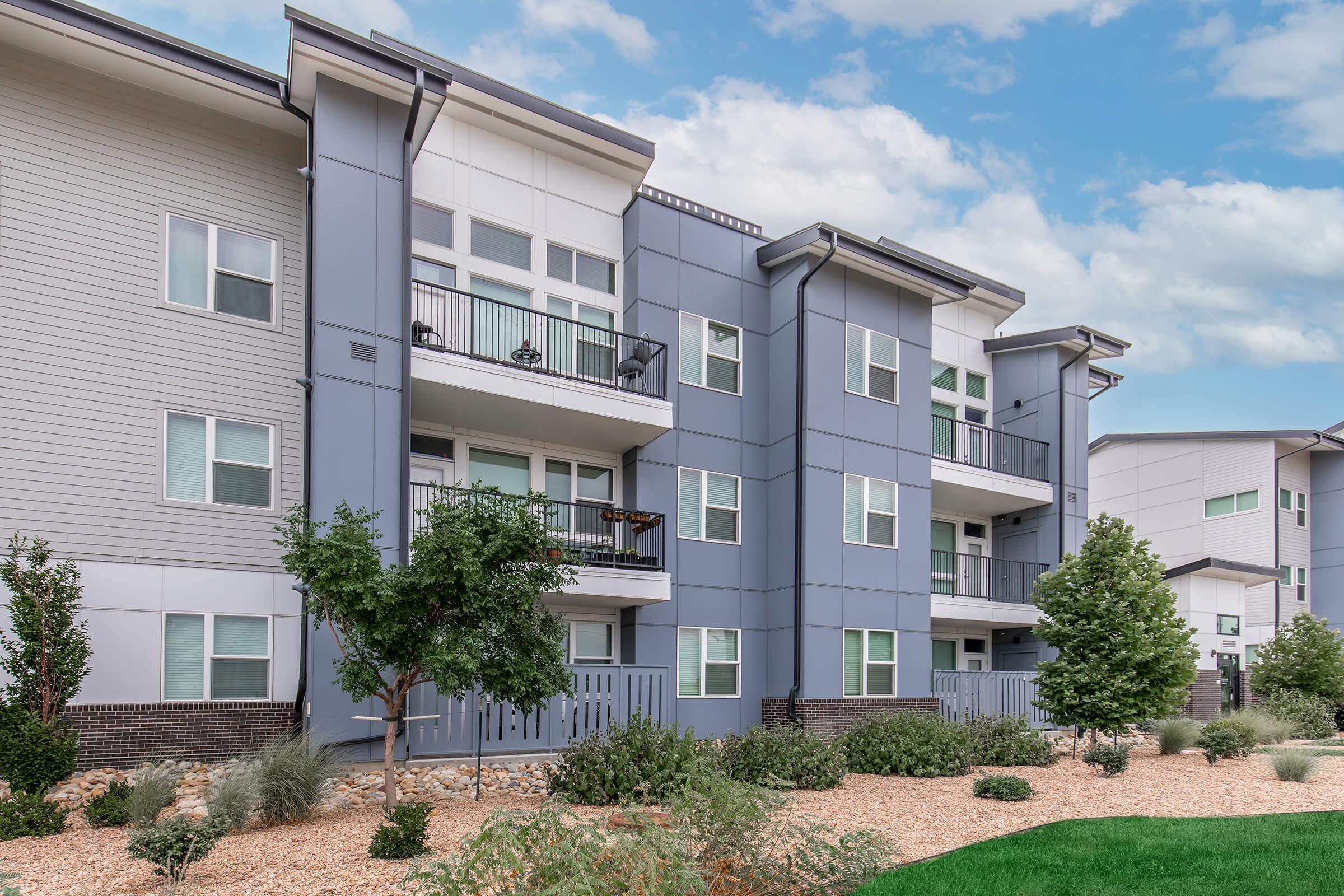 Modern apartment building with three floors, featuring gray and white exterior. Balconies are visible on the upper floors, surrounded by well-maintained landscaping, including small trees and shrubs. The sky is partly cloudy, adding a bright atmosphere to the scene.