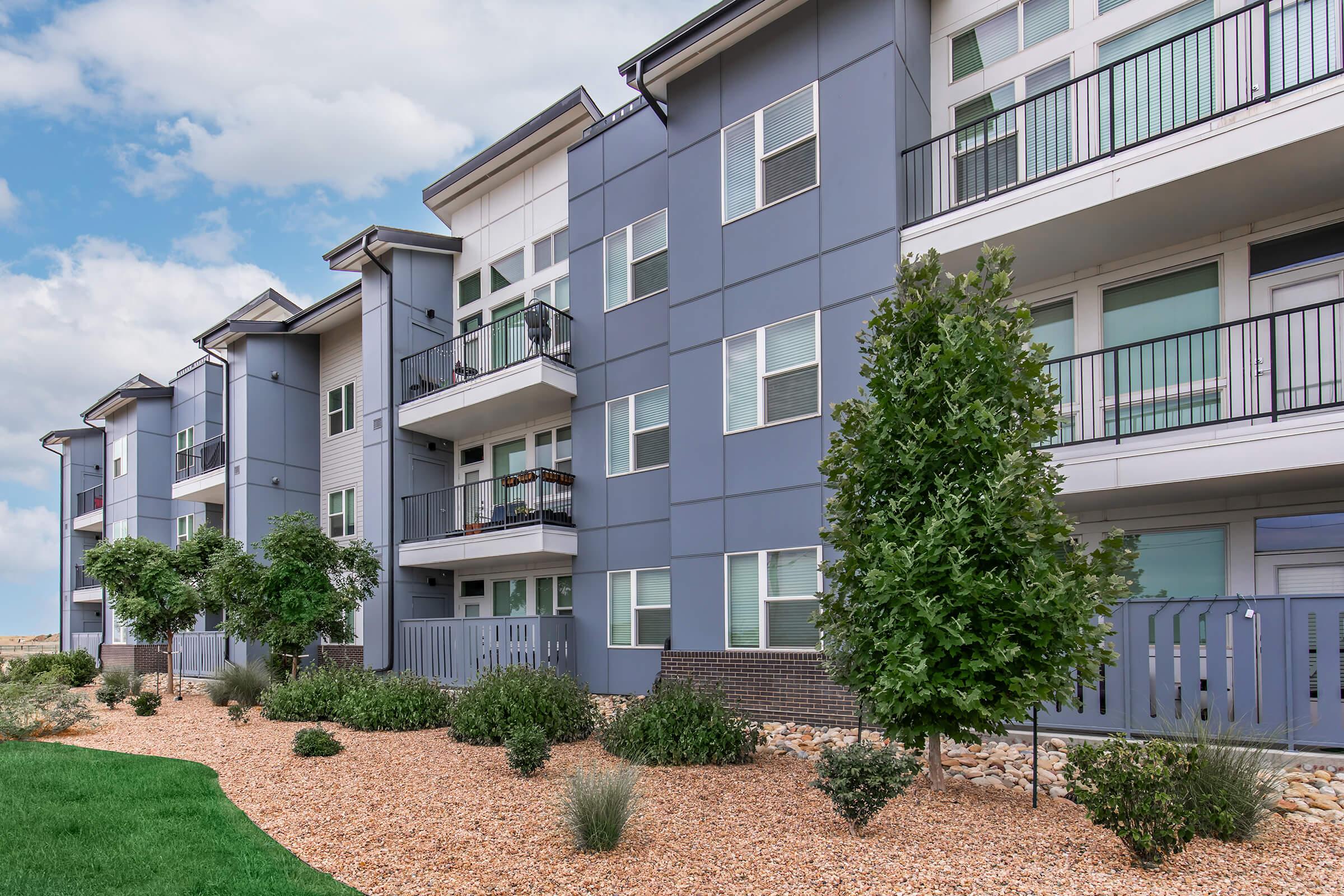 Modern multi-story apartment building with gray exterior, large windows, and balconies. The landscaping features green shrubs and rocks, with a well-maintained lawn in the foreground. The sky is partly cloudy, adding to the inviting atmosphere of the residential area.