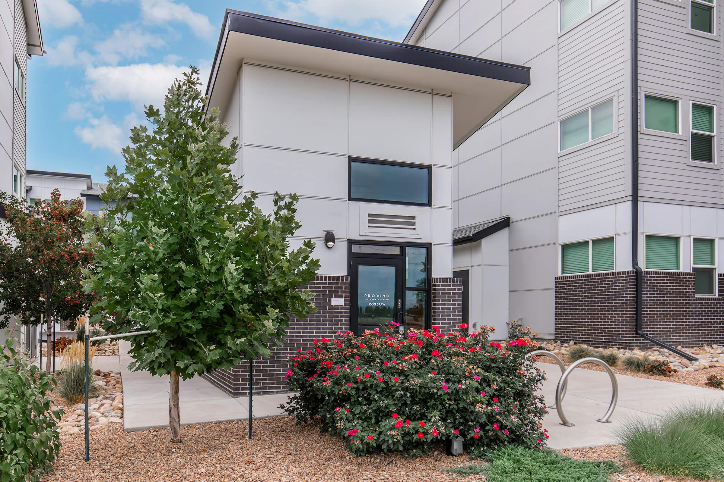 A modern building entrance with large windows, surrounded by neatly landscaped greenery and flowering bushes. The pathway leads to a glass door, featuring a sign, and there's a decorative bike rack nearby. The sky above is partly cloudy, adding a pleasant ambiance to the scene.