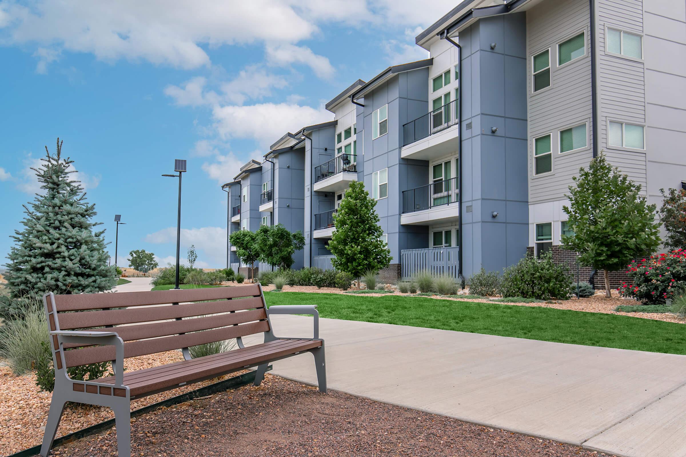 A modern residential building with multiple stories, featuring balconies and large windows. In the foreground, there is a wooden bench along a smooth pathway, surrounded by landscaped greenery, including trees and flowering shrubs, under a partly cloudy sky.