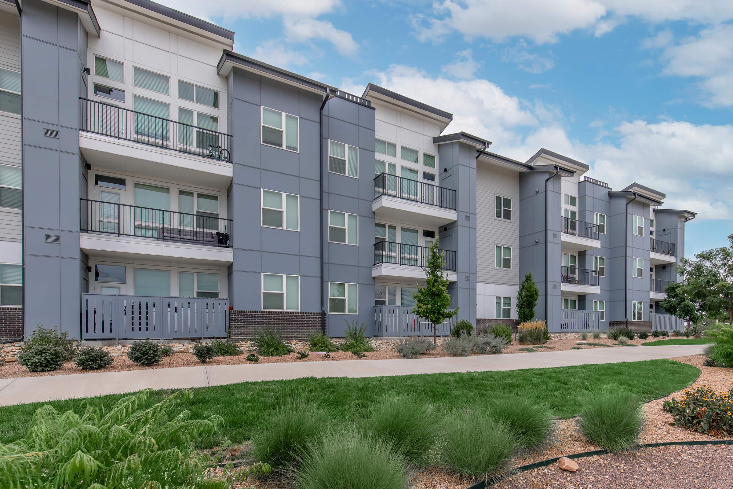 Modern apartment building with multiple units featuring large windows and balconies. The exterior is predominantly gray with some greenery in the foreground, including bushes and ornamental grasses. A paved walkway runs along the front, offering access to the individual units.