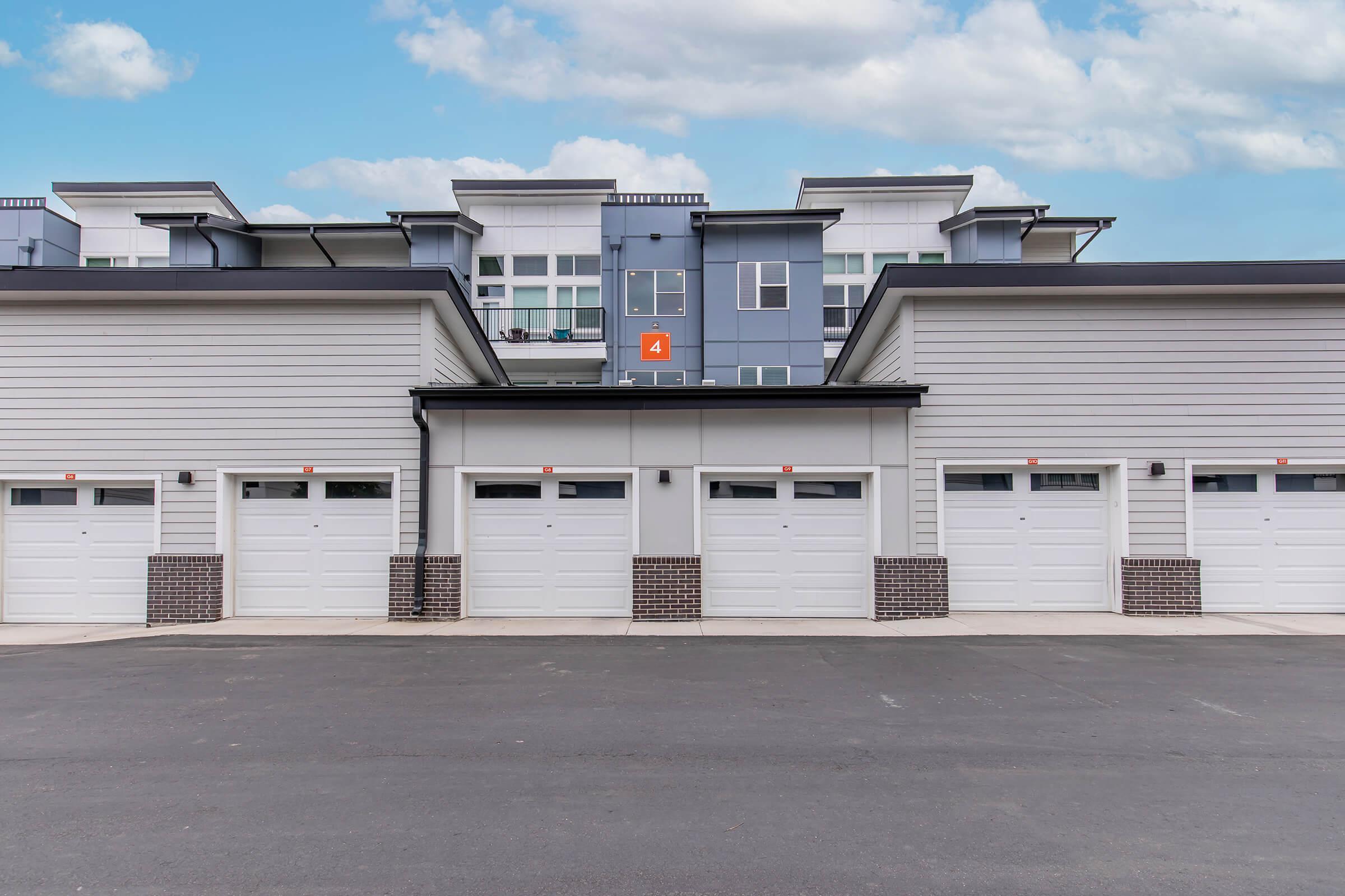A modern residential building featuring several garage doors in a row. The garages are white with small windows and numbers, set against a light gray structure. Above, the building has multiple levels with large windows, providing a contemporary aesthetic against a partly cloudy sky.