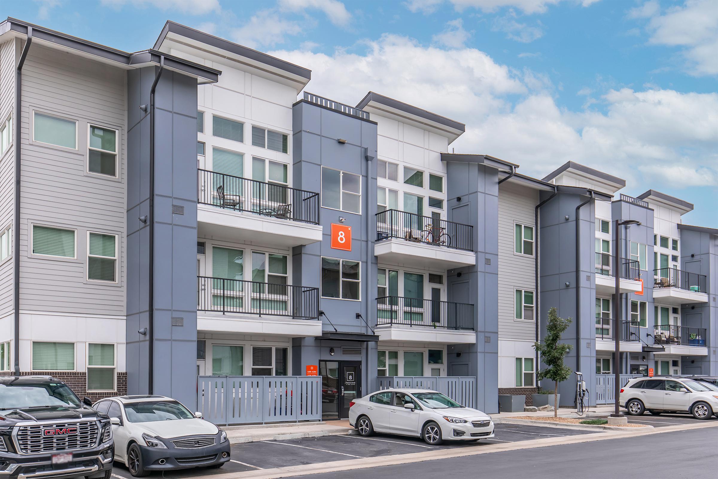 Modern multi-story apartment building with gray and white exterior, featuring balconies and large windows. The entrance is visible with a bright orange number 8 above the door. Several parked cars are in the foreground, and a clear blue sky is in the background.