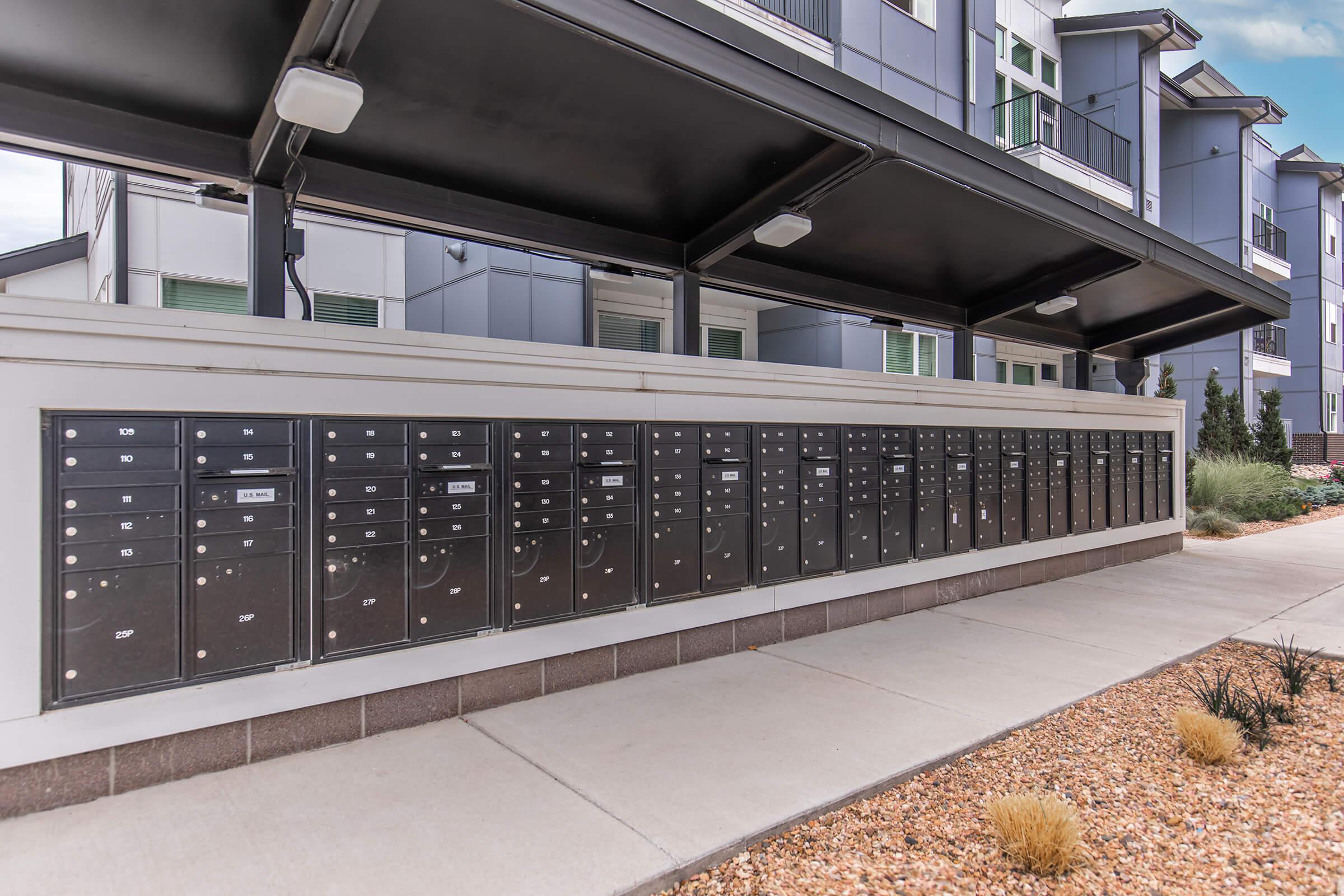 Row of black mailboxes lined up against a modern building, set under a covered awning. Each mailbox has a slot for mail and a number on the front. The pathway in front is paved, with gravel landscaping on either side, providing a contemporary look to the area.