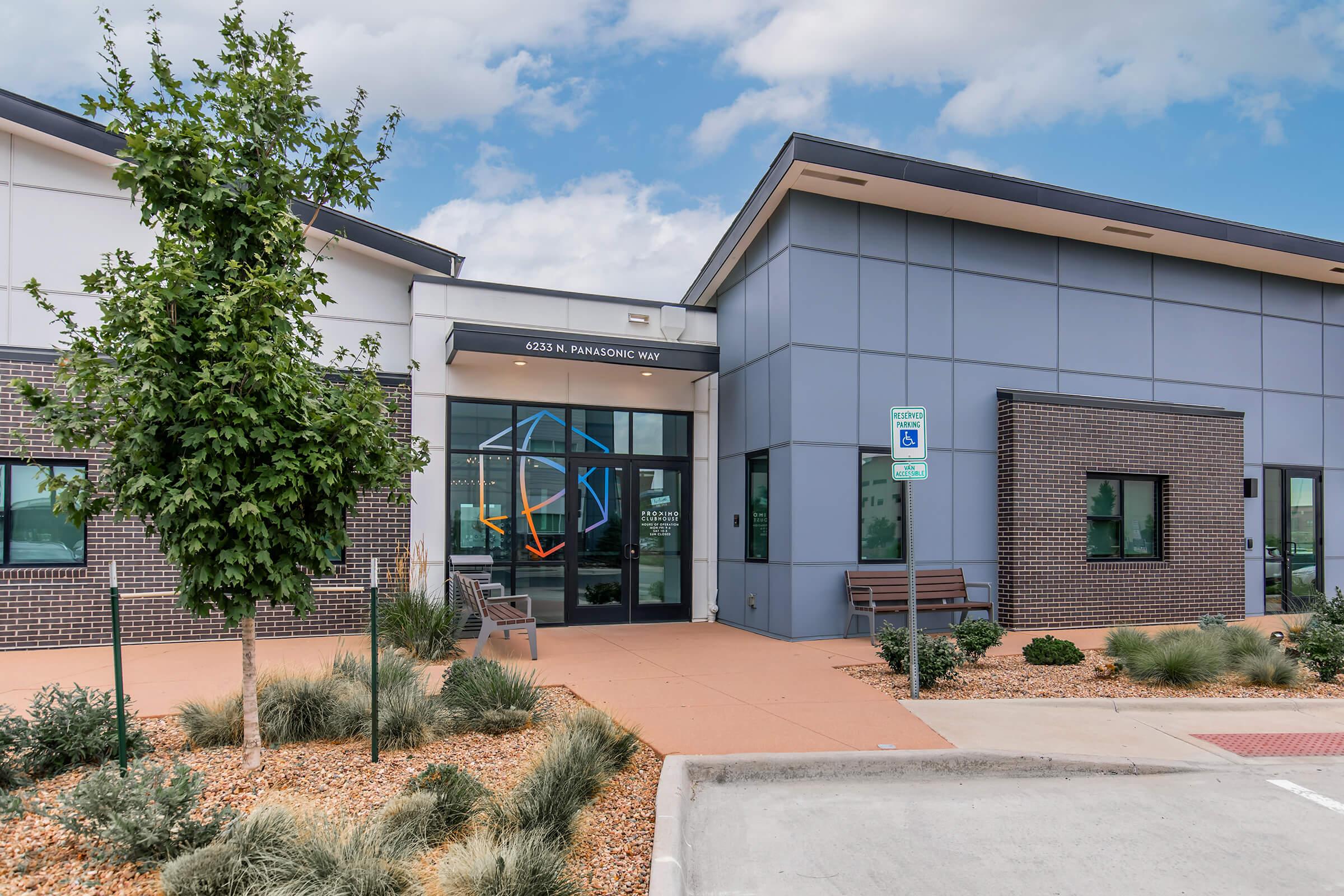 Modern building facade with a large entrance featuring geometric designs on the glass doors. The exterior includes a mix of gray and brick materials, landscaped with small shrubs and gravel. There are visible benches and a parking area with accessible parking signage. Cloudy sky overhead.