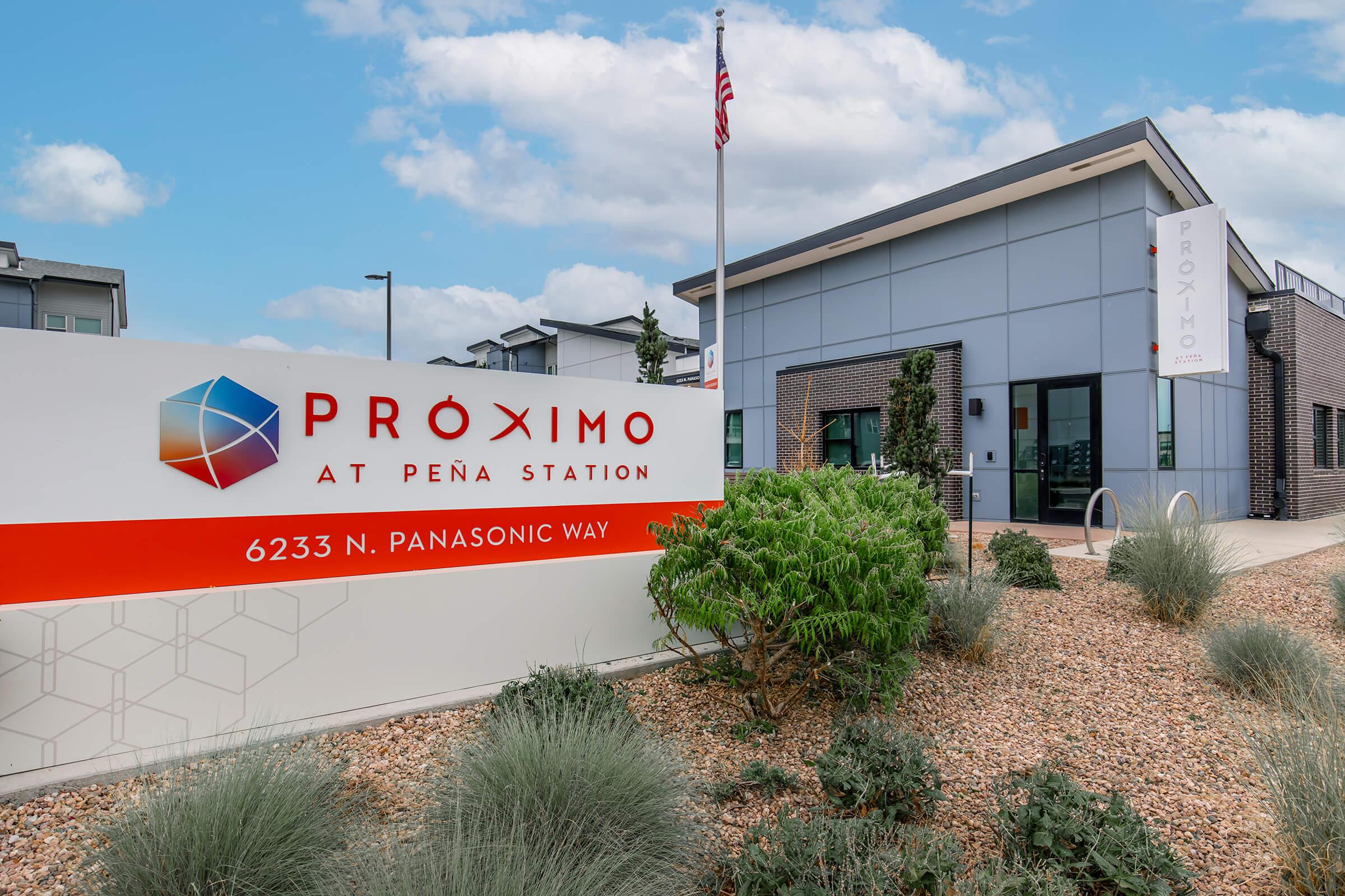 Sign for "Próximo at Peña Station" displayed prominently in front of a modern building. The sign includes the address "6233 N. Panasonic Way" and is accompanied by greenery and landscaped features. The building showcases a contemporary design with large windows and a welcoming entrance.