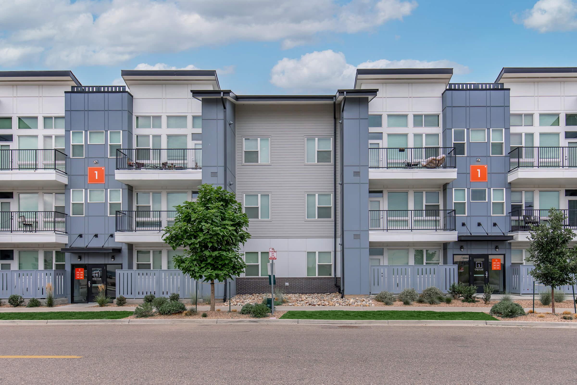 Modern apartment building with a symmetrical facade featuring multiple balconies and large windows. The building is designed with a combination of gray and white exterior elements. There are several trees and landscaped areas in front, along with street signage. The sky is partly cloudy.
