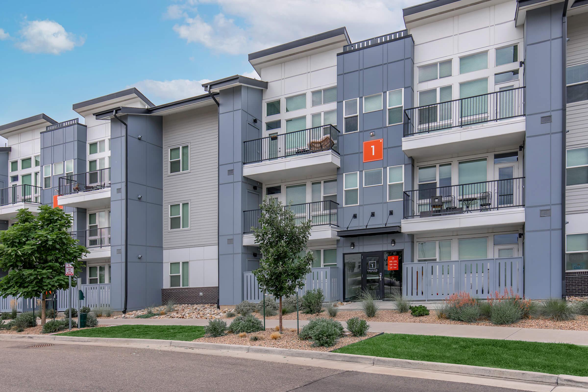 Modern apartment building with multiple stories and balconies. The exterior features a mix of gray and white panels with large windows. A prominent orange number "1" is displayed near the entrance. The landscaped area includes small trees and shrubs, and the scene is set on a wide street.
