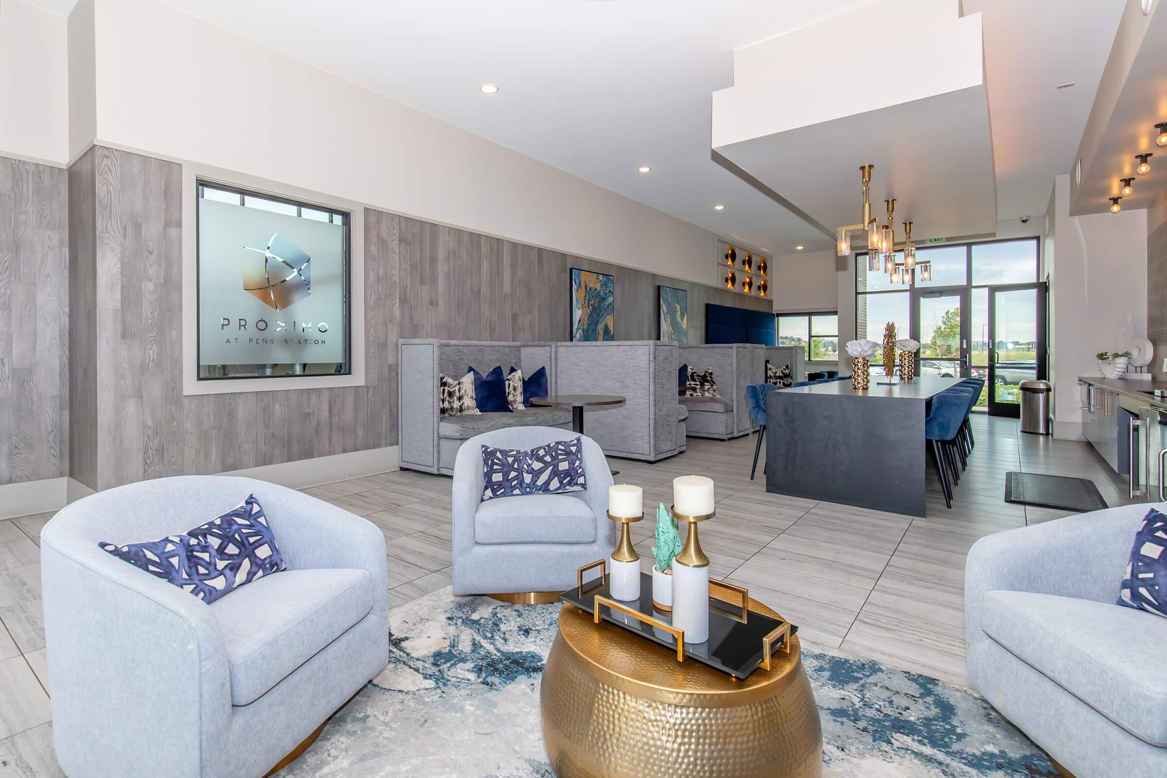 Modern interior of a stylish lounge area featuring light blue armchairs, a round gold coffee table, and a large TV display on the wall. The room has elegant lighting fixtures, wooden accents, and a mix of decorative cushions. Large windows allow natural light to brighten the space.