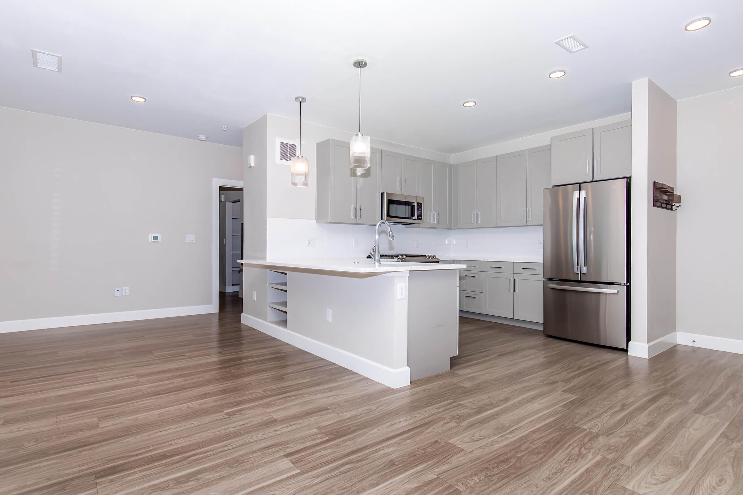 Modern kitchen featuring light gray cabinets, stainless steel appliances, and a large island with seating. The space has hardwood-style flooring and is well-lit with recessed lighting and pendant lights above the island. It has an open layout with a spacious, minimalist design.