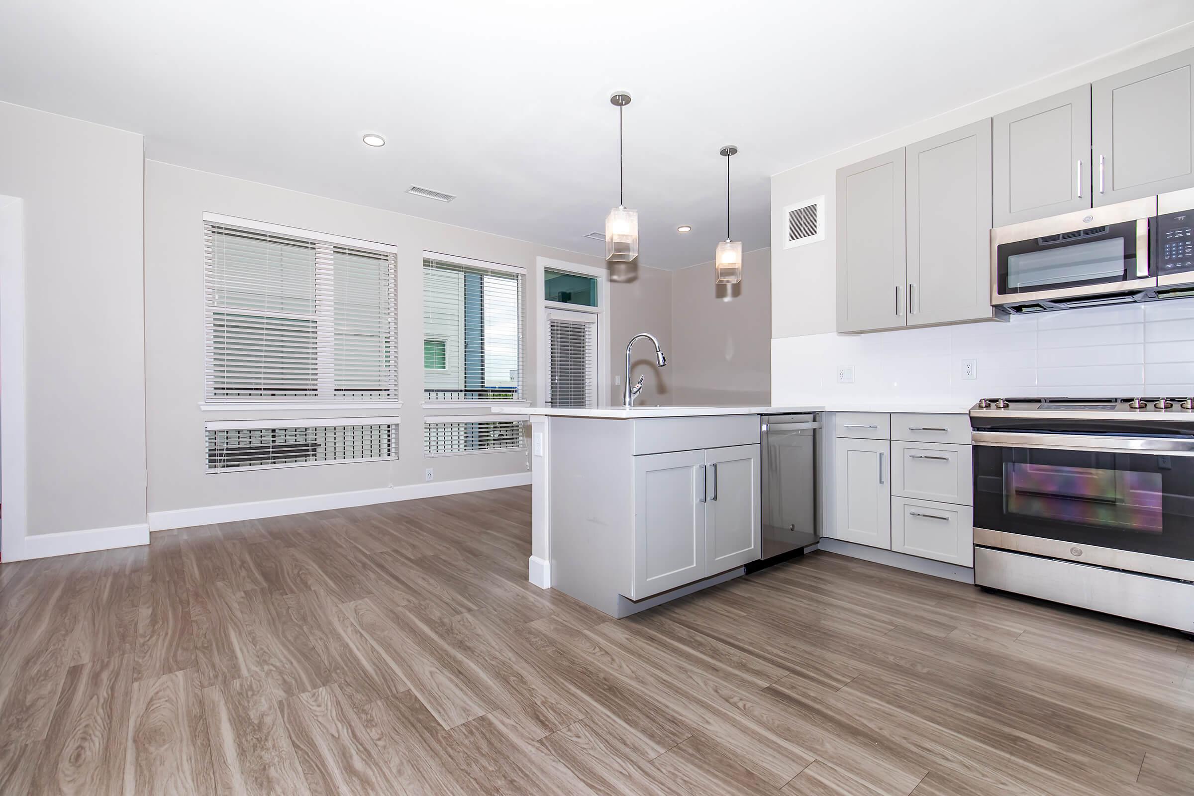 Modern kitchen with light gray cabinets, stainless steel appliances, and a large island. Features include pendant lighting and a spacious layout with hardwood flooring. Large windows let in natural light, enhancing the open and inviting atmosphere.