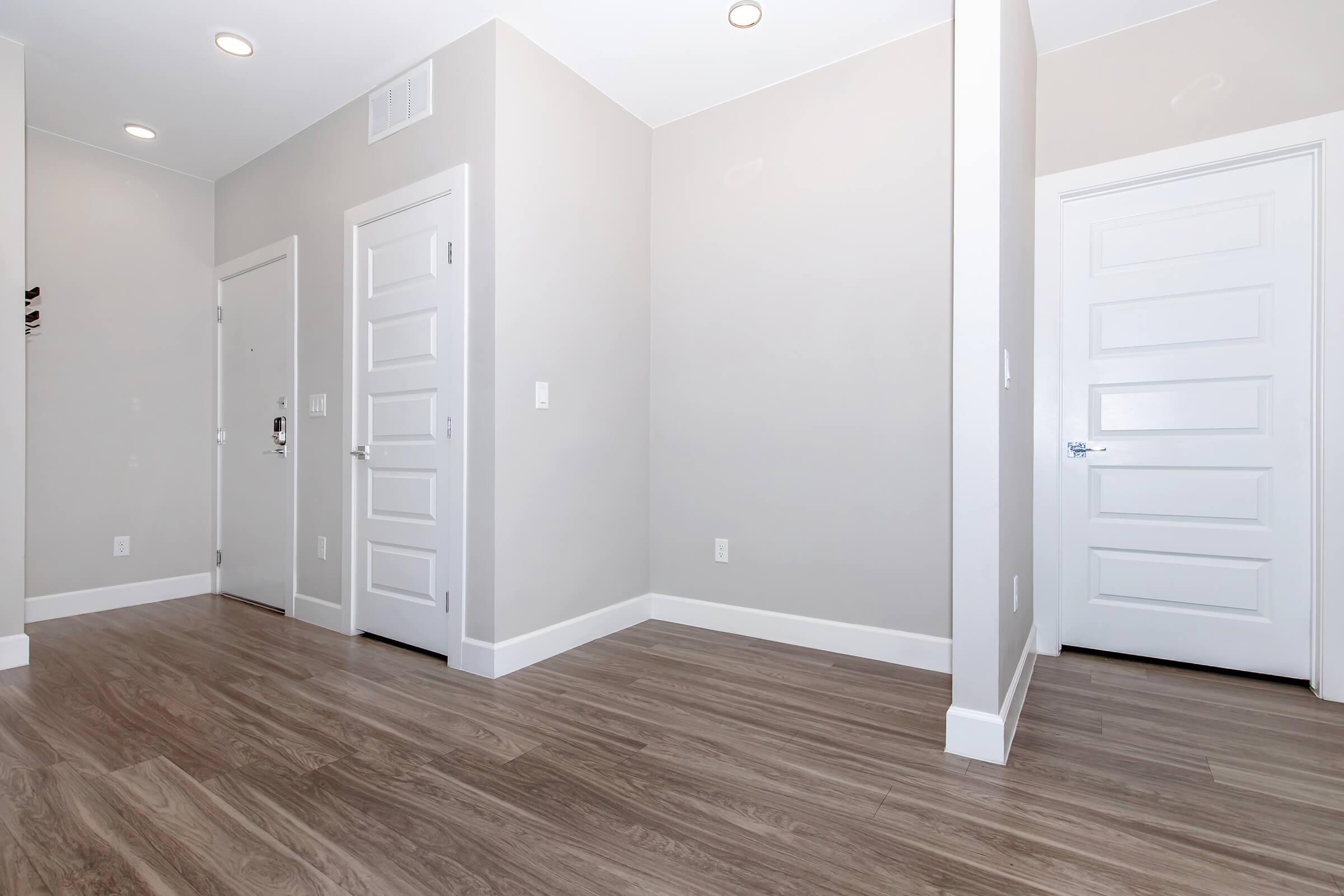A modern hallway featuring neutral-colored walls and light wood flooring. The space includes multiple doors, one leading to an entryway, with minimalistic white trim and a ceiling with recessed lighting. The overall aesthetic is clean and spacious, reflecting contemporary design.