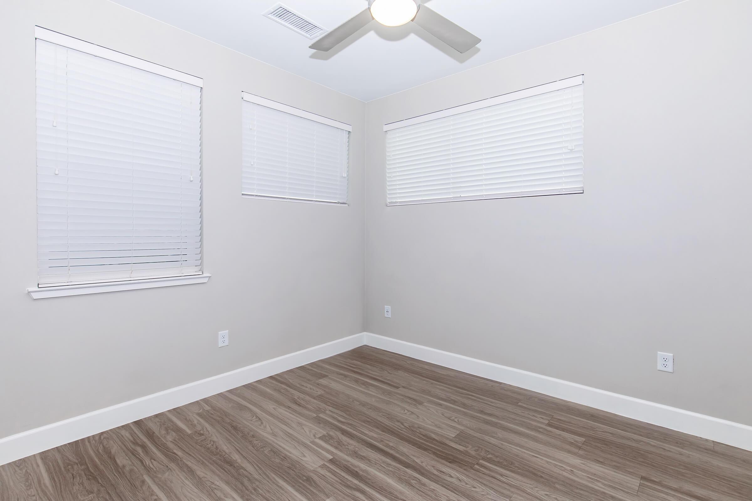 An empty room with light-colored walls and two windows featuring white blinds. The flooring is a light wood design, and there is a ceiling fan overhead. The space is devoid of furniture or decorations, creating a minimalist and spacious feel.