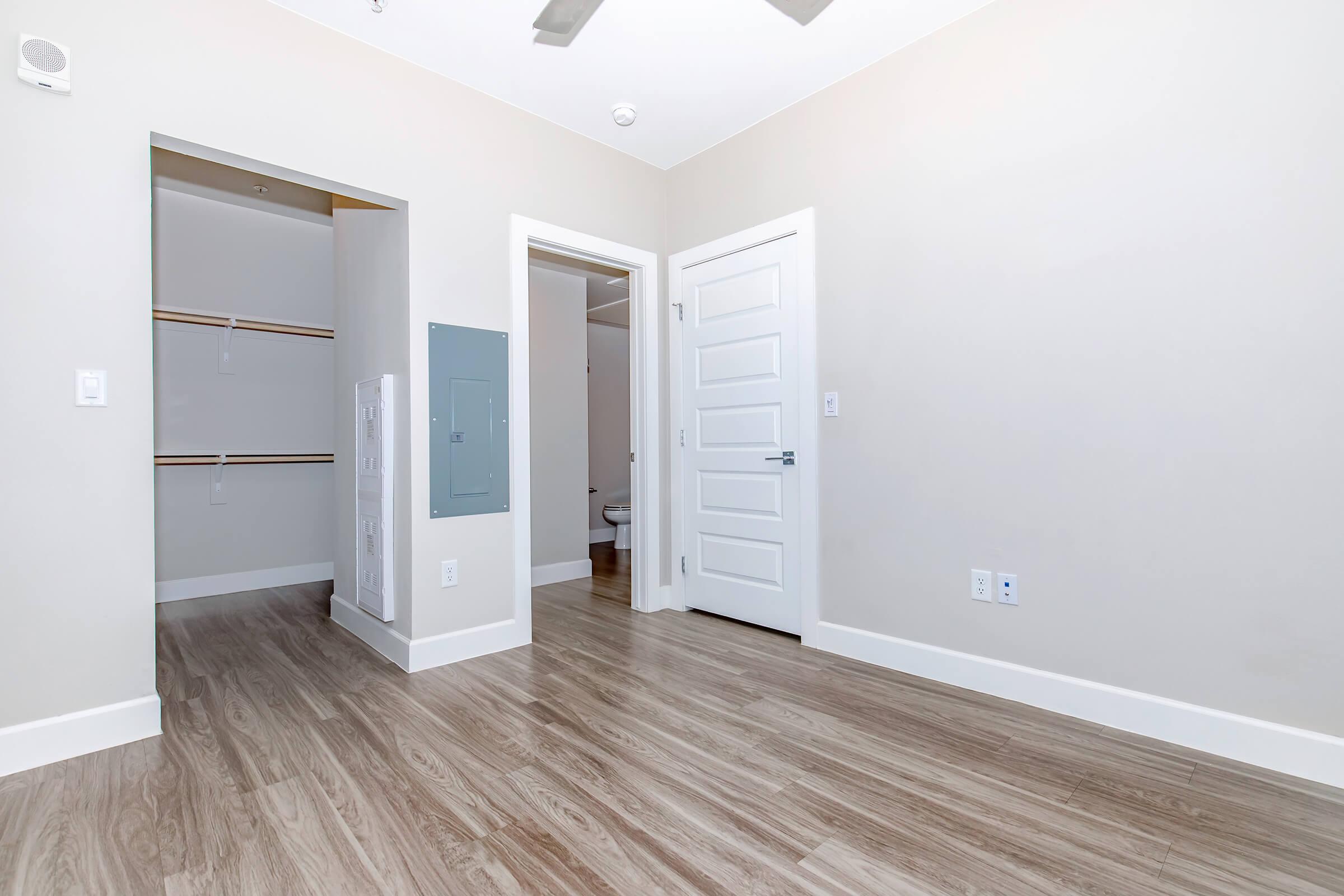 A spacious, minimally furnished room with light-colored walls and wooden flooring. There is a closet with shelving on the left, a doorway leading to a bathroom, and a white door on the right. A ceiling fan is visible, and the overall ambiance is bright and modern.