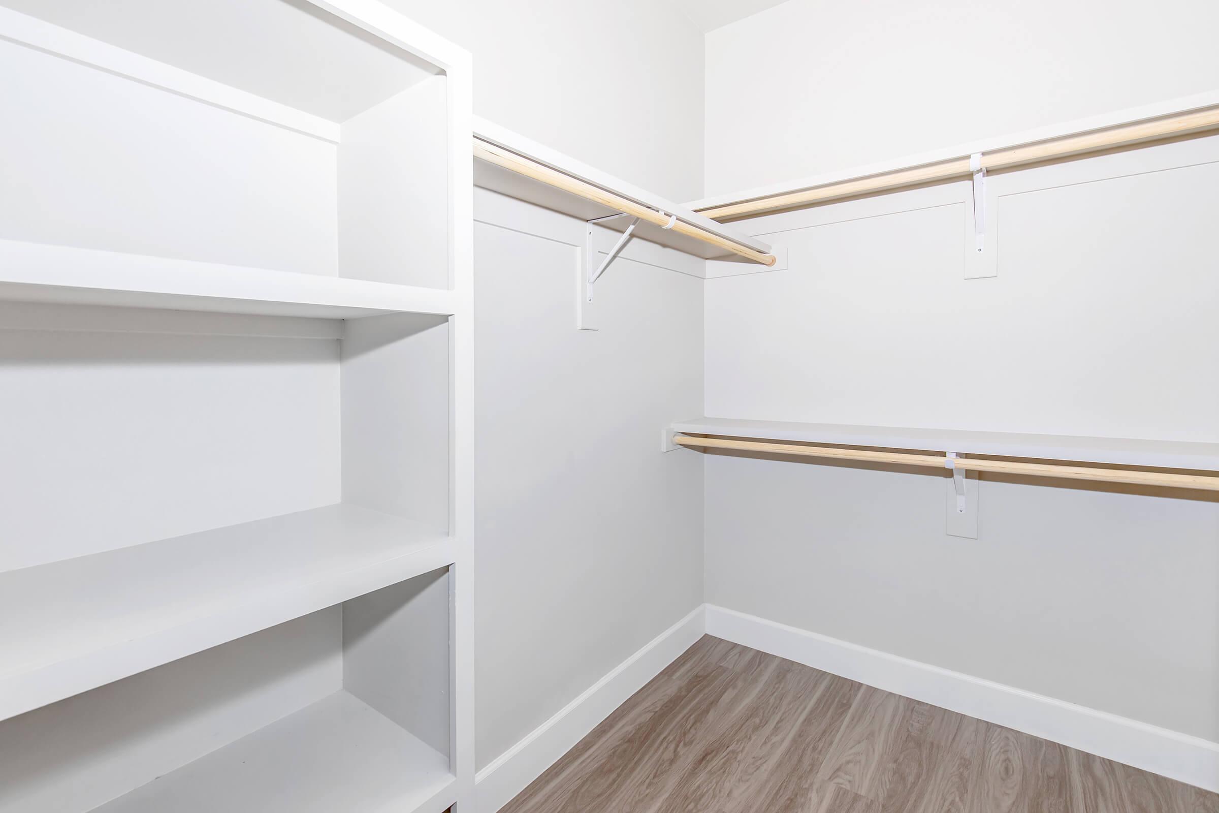 A minimalist closet interior featuring white shelves and two hanging rods on smooth white walls. The floor is light wood, creating a clean and bright space suitable for organizing clothes and accessories.