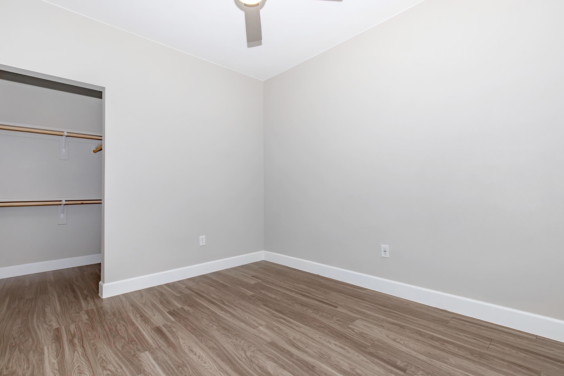 A spacious, empty room with light gray walls and wooden flooring. There is a small closet to the left with wooden shelves. A ceiling fan is visible, and the room is well-lit, creating a clean and bright atmosphere.