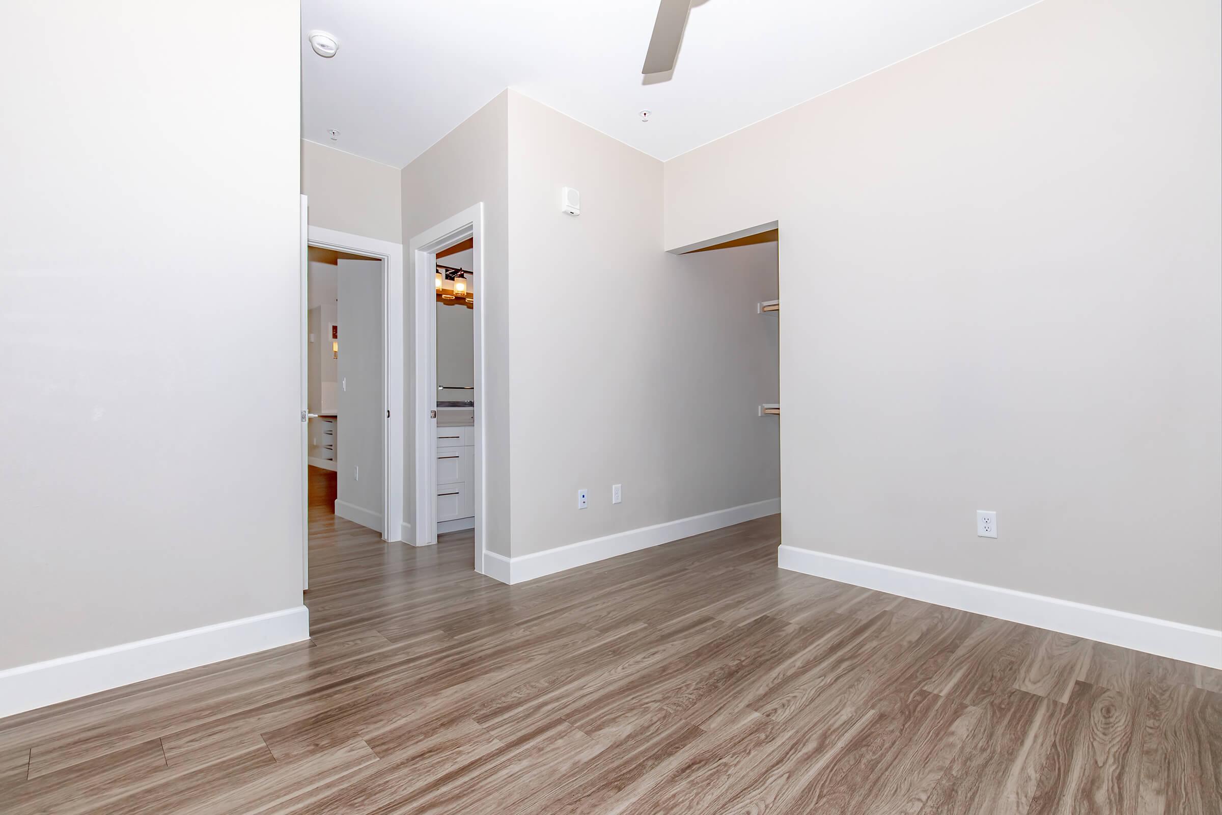 A clean, modern room with light-colored walls and hardwood flooring. A doorway leads to another room and a small closet is visible on the right. The ceiling has a ceiling fan, and there is ample natural light, creating a spacious and airy atmosphere.