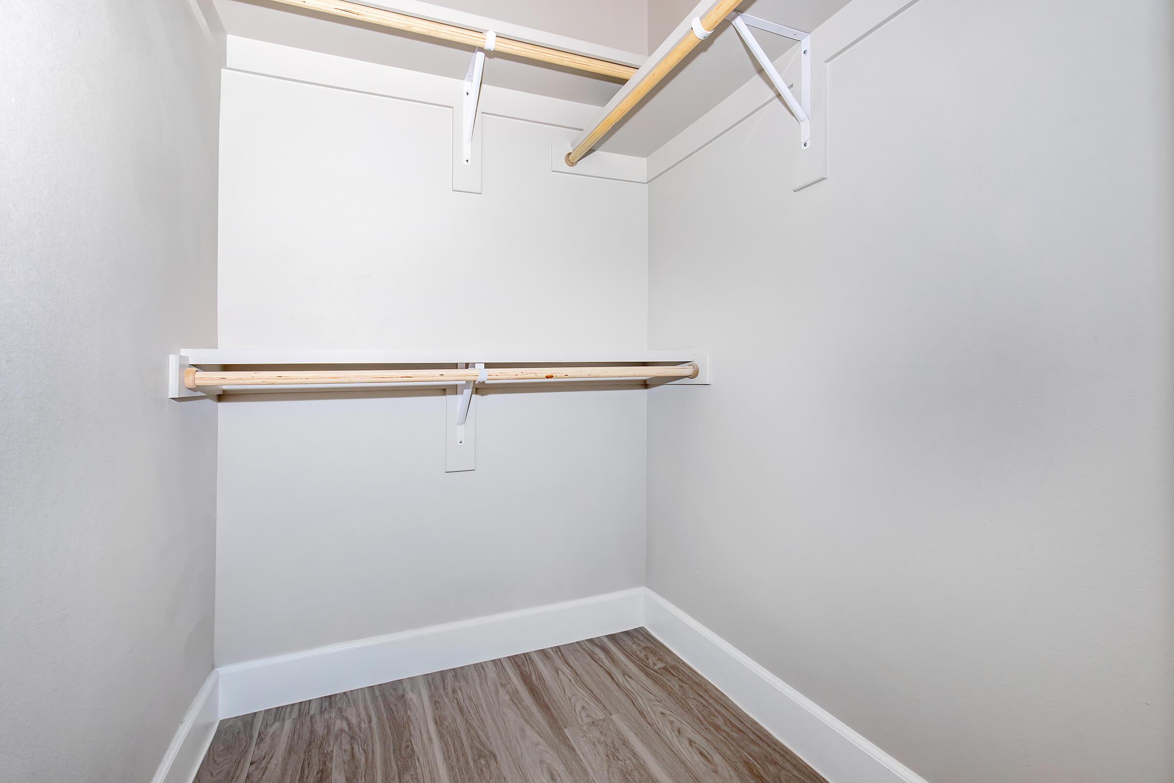 A spacious and empty closet with two wooden rods for hanging clothes, featuring light-colored walls and a wooden floor. The minimalistic design emphasizes the uncluttered space, making it suitable for organizing items or storage.