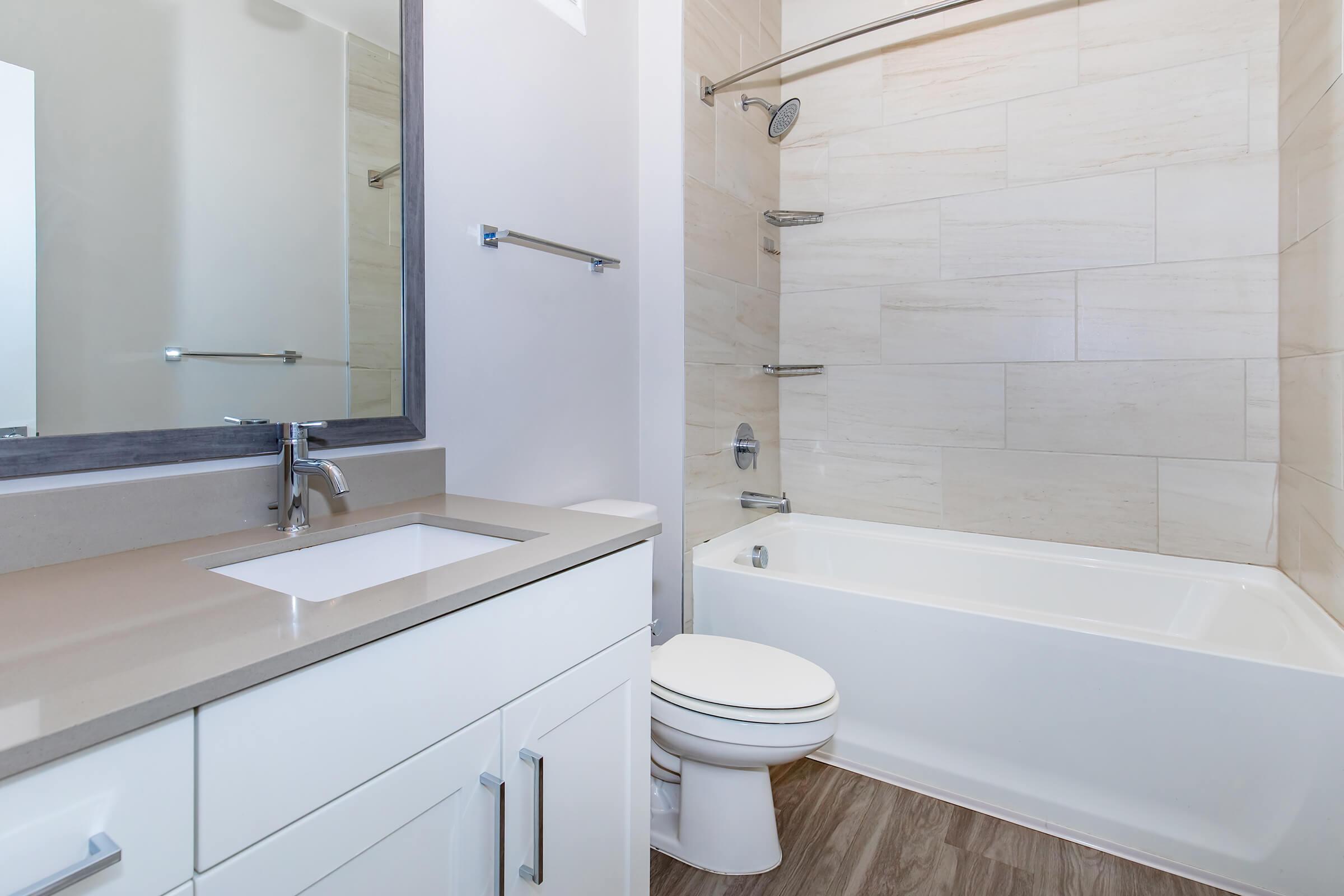 Modern bathroom interior featuring a bathtub with a shower, a white toilet, and a sleek vanity with a sink. The walls are lined with light-colored tiles, and there is a large mirror above the sink. Natural light floods the space, enhancing the clean and contemporary design.