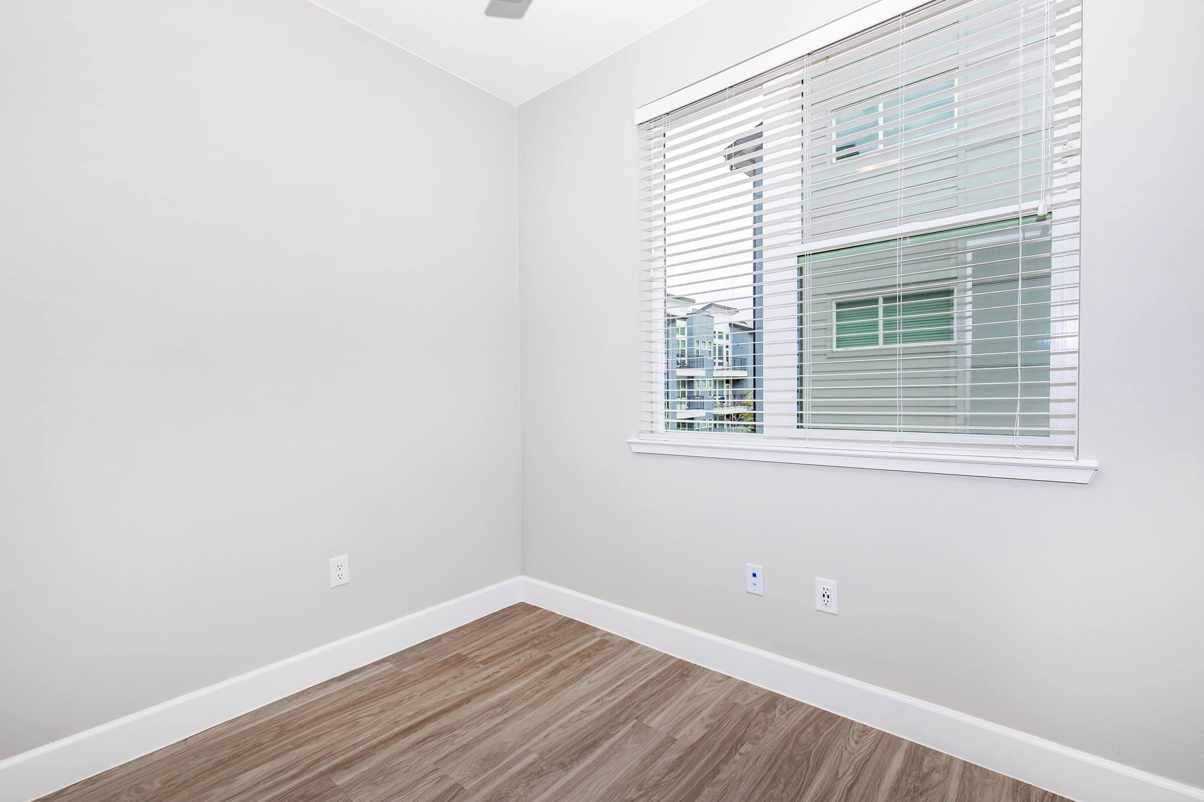 A corner of a room with light gray walls and a light brown laminate floor. A window with white blinds allows natural light to enter, revealing a view of an exterior with greenery. The room appears empty and unfurnished, creating a bright and inviting atmosphere.