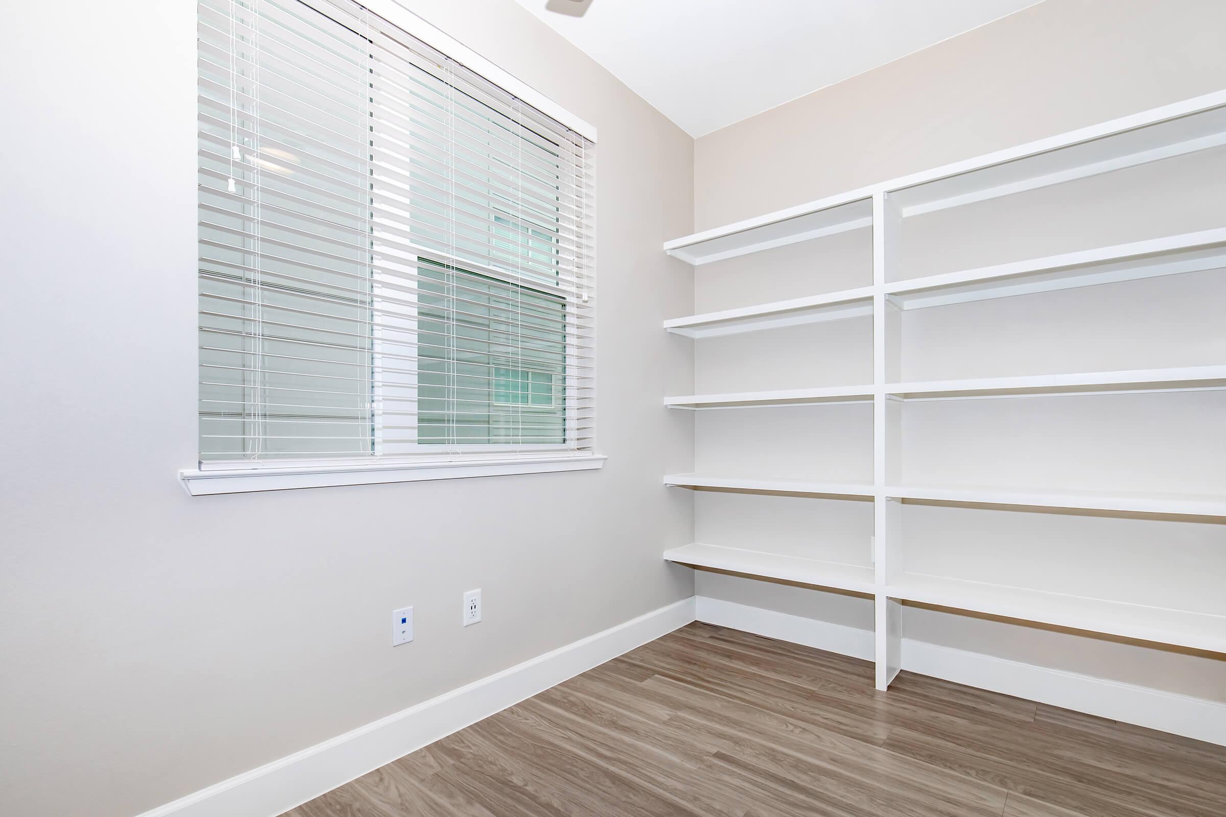 A light, empty room with beige walls and a window covered with blinds. There are white built-in shelves against one wall and a smooth wooden floor. The space is well-lit and minimalistic, ideal for storage or as an office area.