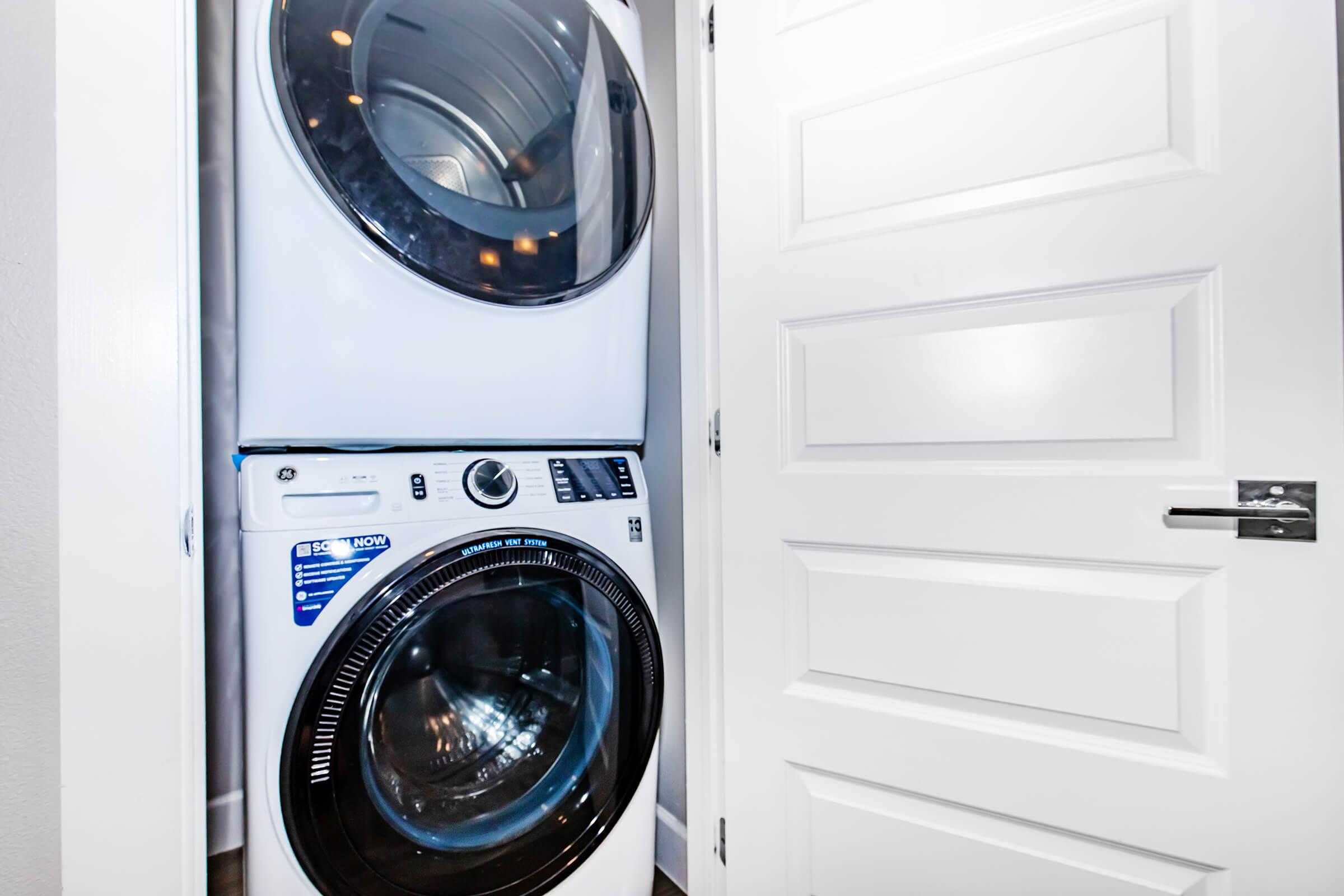 A modern stacked washer and dryer unit located in a small laundry space beside a closed white door. The appliances are sleek and offer a contemporary look, with a focus on efficiency and design. The room is well-lit, enhancing the clean lines of the appliances.