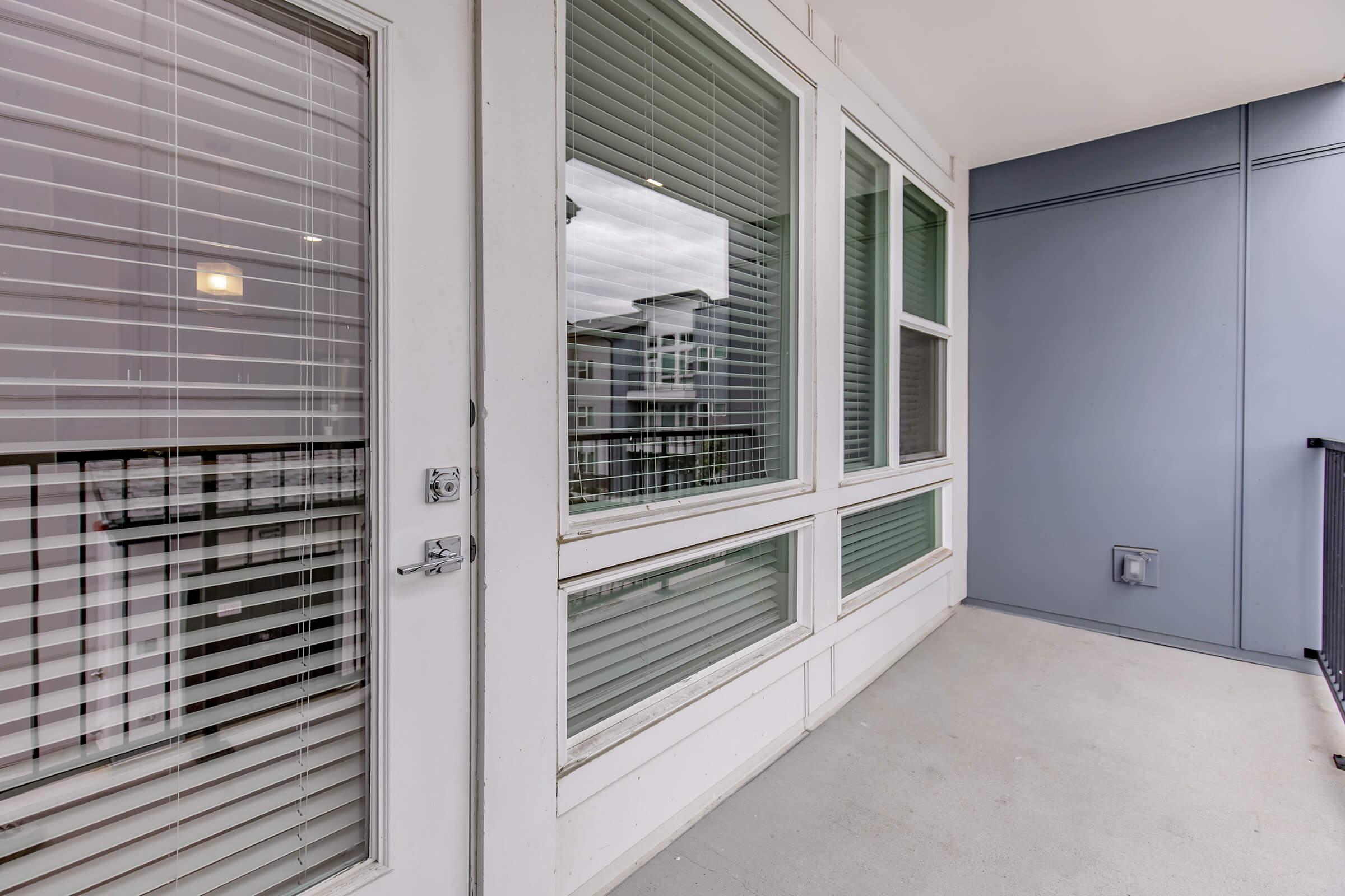 A modern balcony with large windows and sliding doors, showcasing a clean exterior design. The view is partially obscured by blinds, and the floor is plain concrete. The surrounding architecture includes multiple levels of buildings visible through the glass.