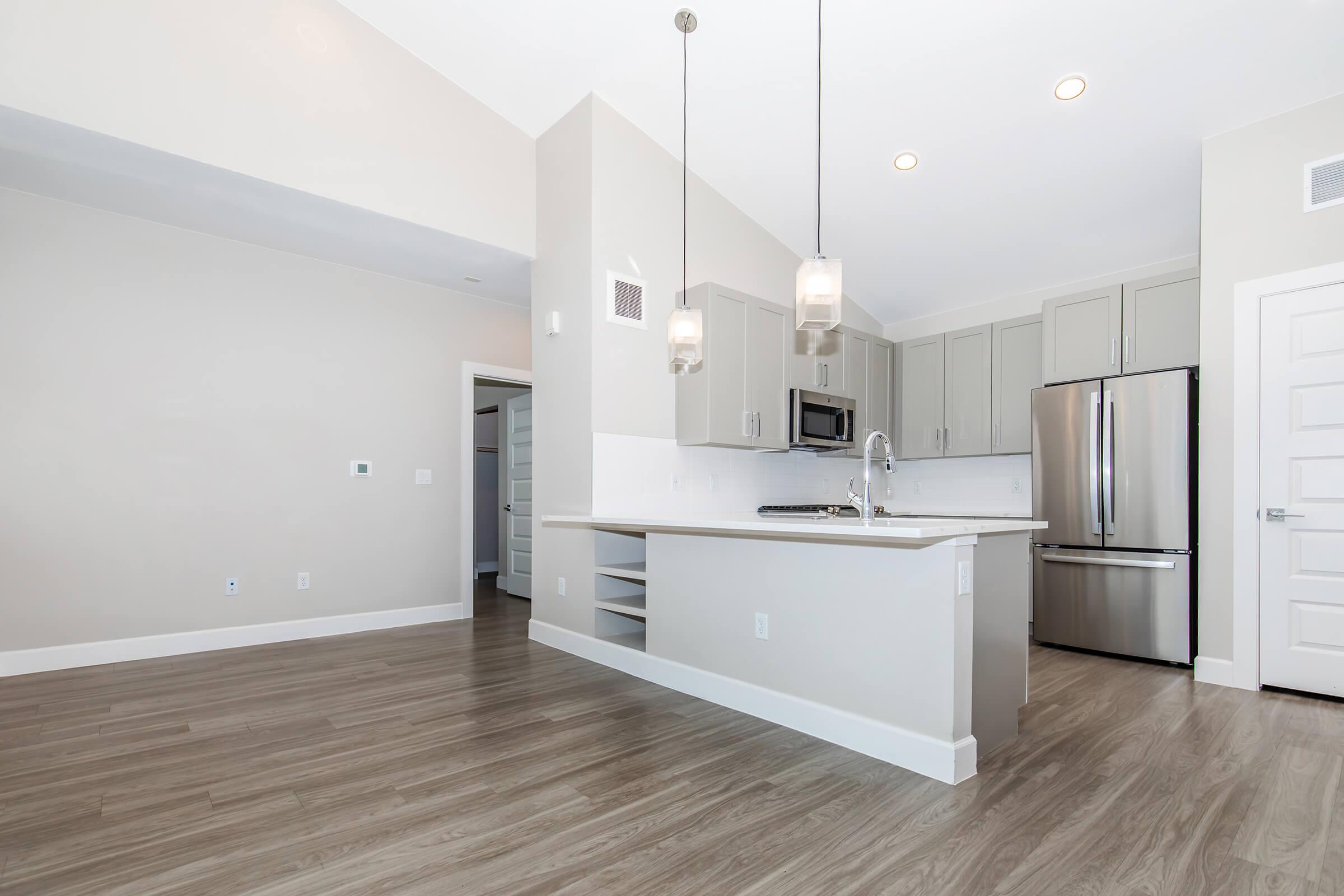 A modern kitchen with light-colored walls and wood-style flooring. It features gray cabinetry, stainless steel appliances including a refrigerator and microwave, and pendant lighting above the island. There is an open layout with a doorway leading to another room. Bright and spacious atmosphere.