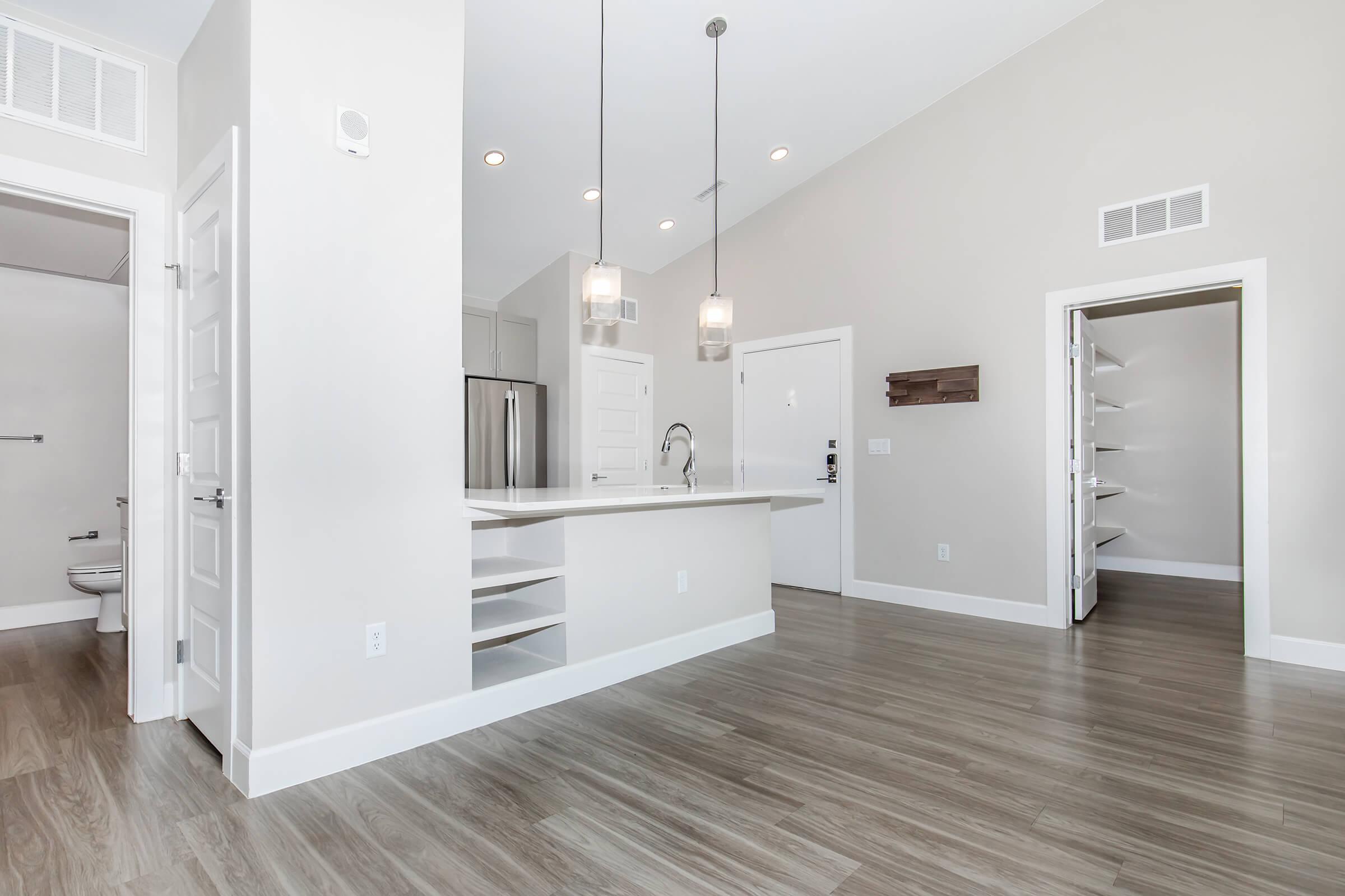 A spacious and modern open-concept living area featuring light-colored walls and hardwood flooring. The kitchen includes a breakfast bar with pendant lighting, and there are doors leading to a pantry and a bathroom. Natural light fills the space from large windows, creating a bright and inviting atmosphere.