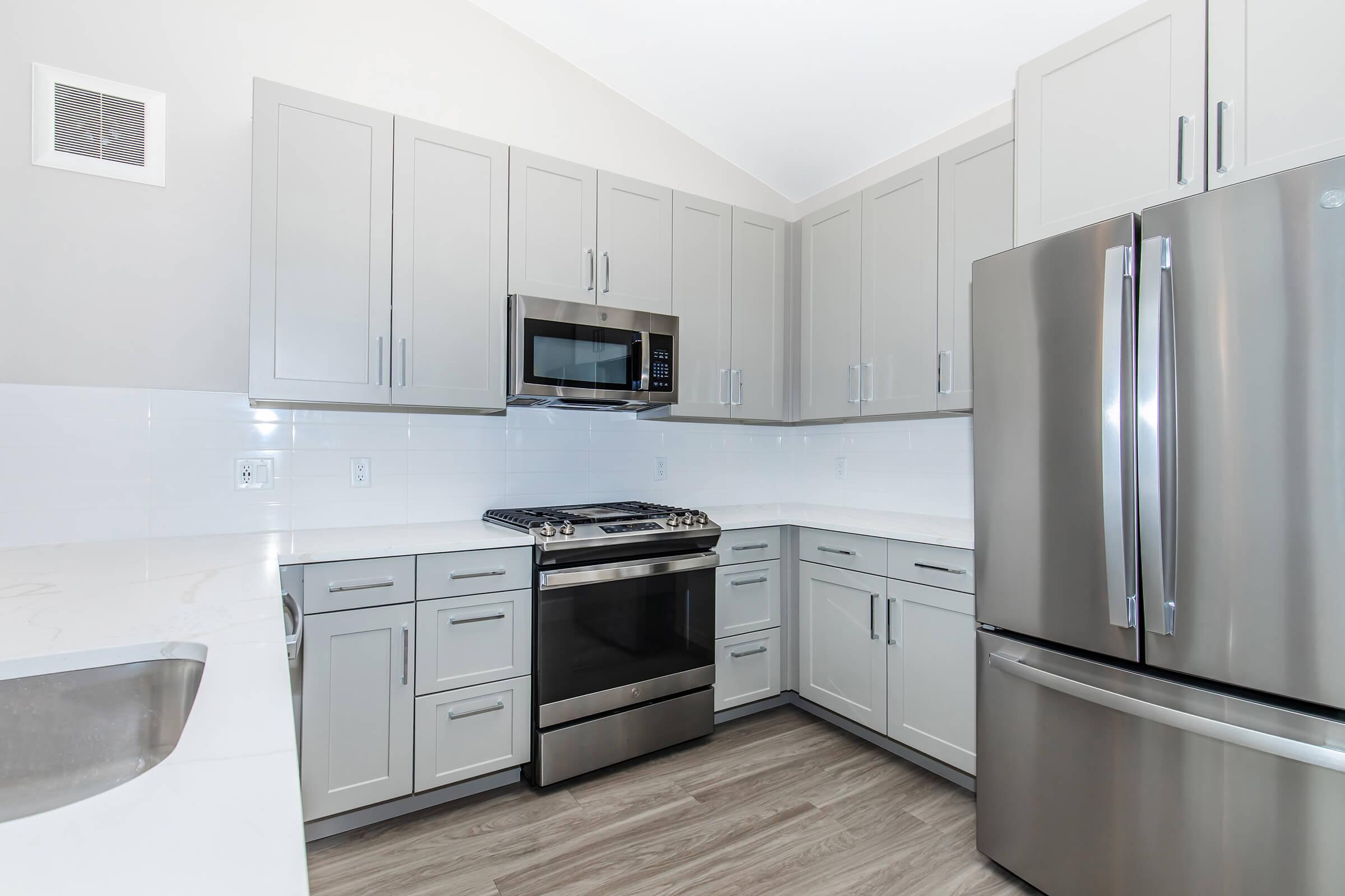 Modern kitchen featuring light gray cabinets, stainless steel appliances including a microwave, oven, and refrigerator. The countertop is made of a light material, and the floor has a wood-like finish. The kitchen has an open layout with ample storage and a bright, clean appearance.