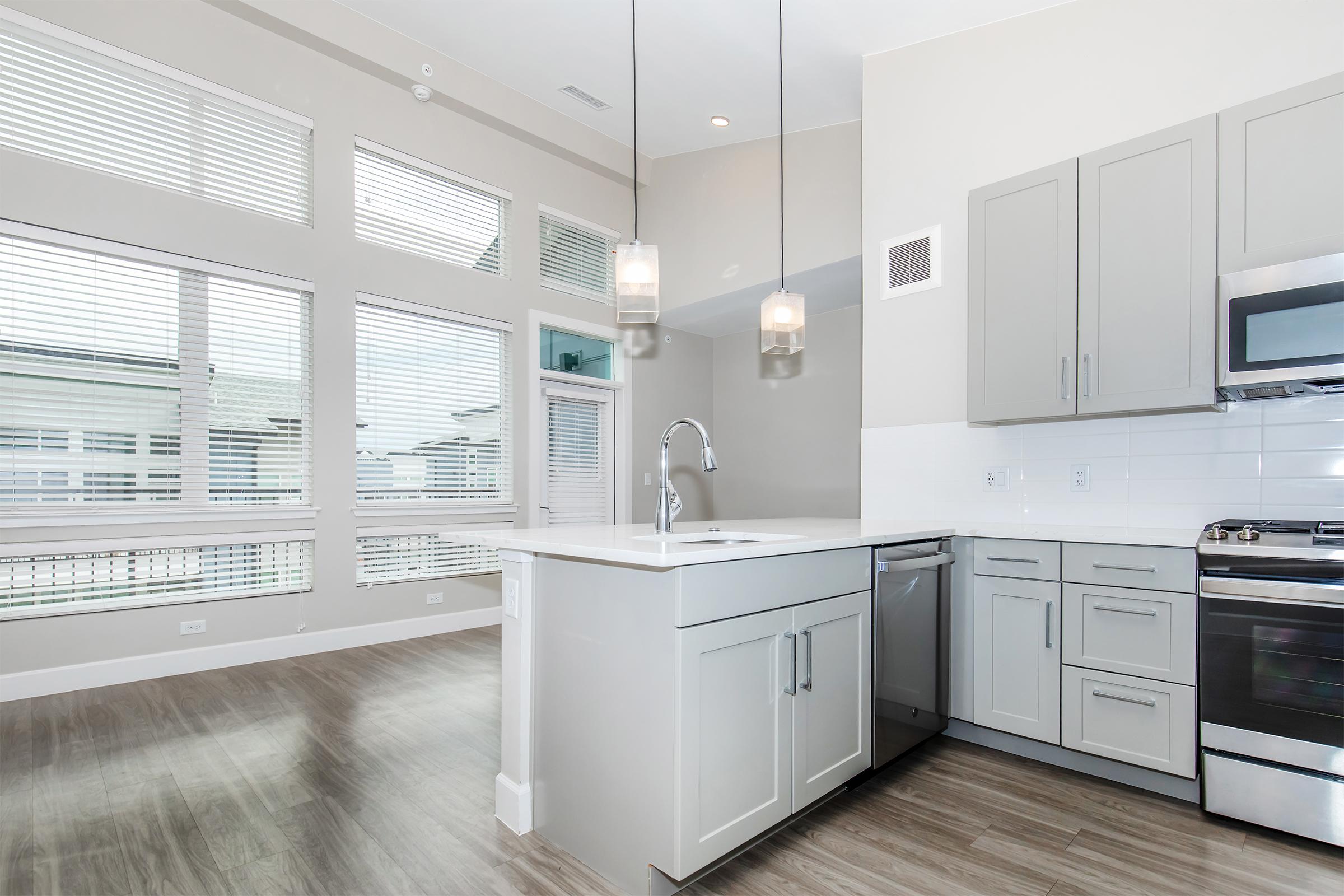 Modern kitchen with light gray cabinetry, stainless steel appliances, and a large island with a sink. The space features large windows allowing natural light to fill the room, highlighting the wooden floor and neutral-colored walls. Pendant lights hang above the island, enhancing the contemporary design.