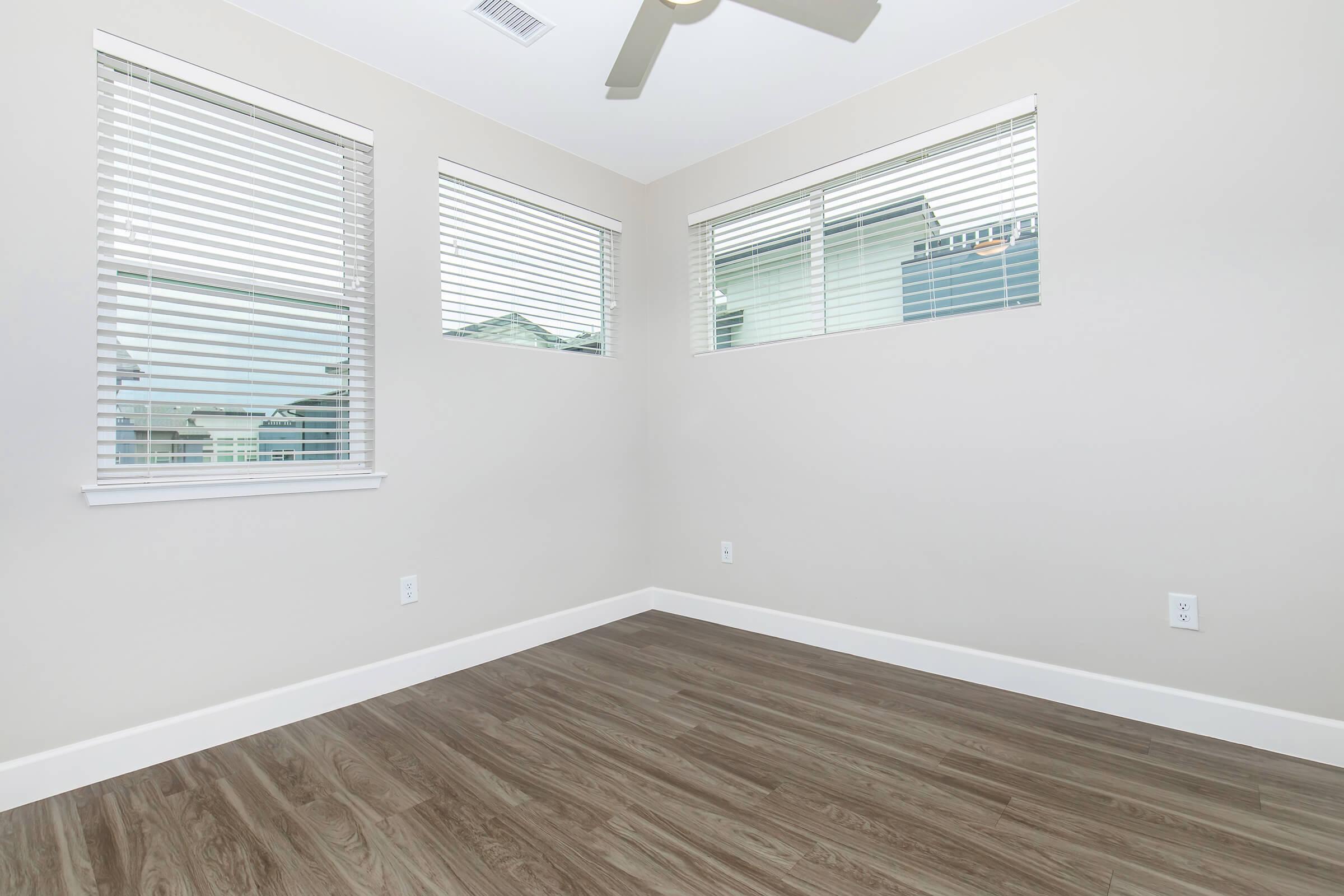 A bright, empty room with light-colored walls and large windows featuring white blinds. The floor is finished with wood-like laminate, and a ceiling fan is mounted above. There are no furnishings, emphasizing the spaciousness of the area.
