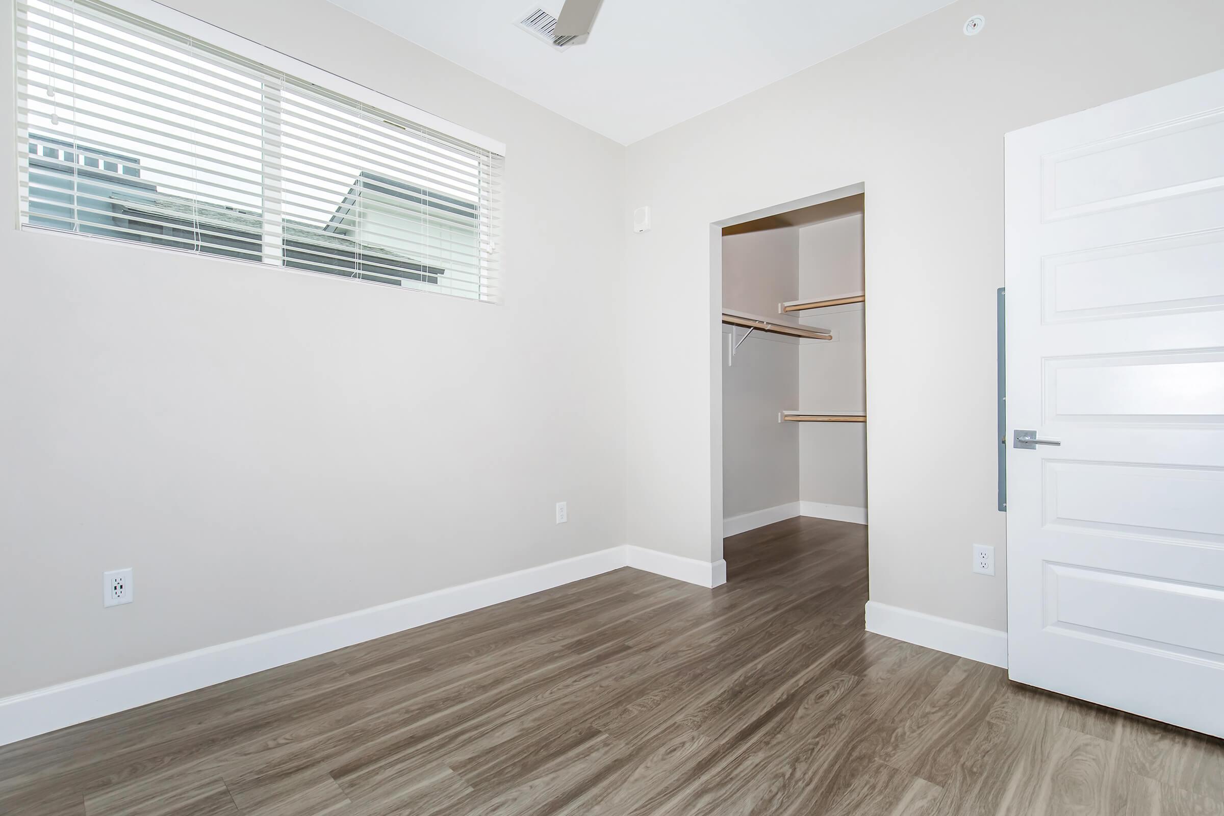 A spacious, empty room featuring light-colored walls and a window with horizontal blinds. The floor is made of brown laminate. A doorway on the right leads to a closet with open shelving. The overall ambiance is bright and minimalistic.