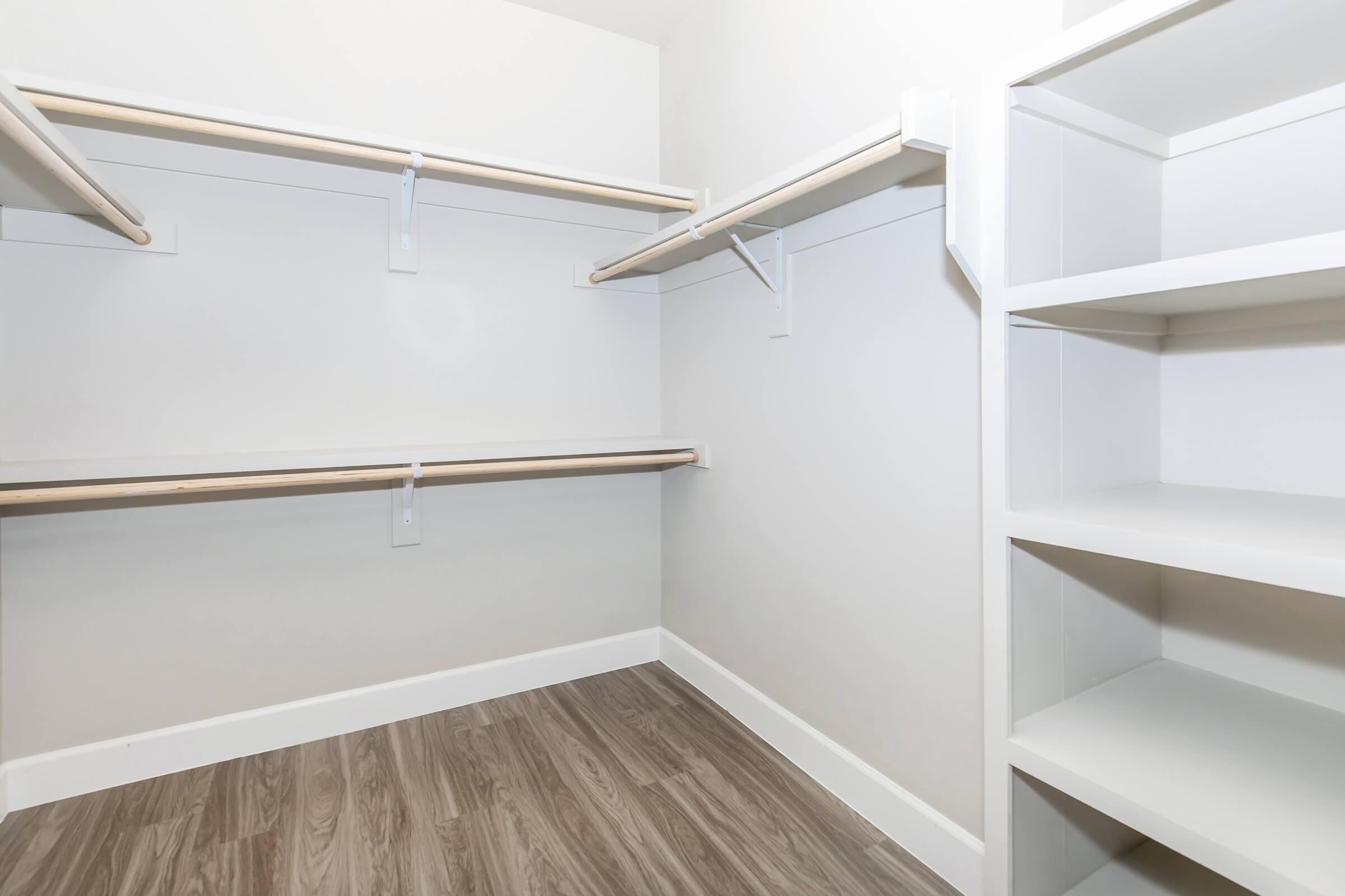 A spacious, empty closet featuring white walls, wooden shelves on either side, and a light brown laminate floor. The shelves are organized with brackets, and there are no items stored inside, giving a clean and minimalist appearance.