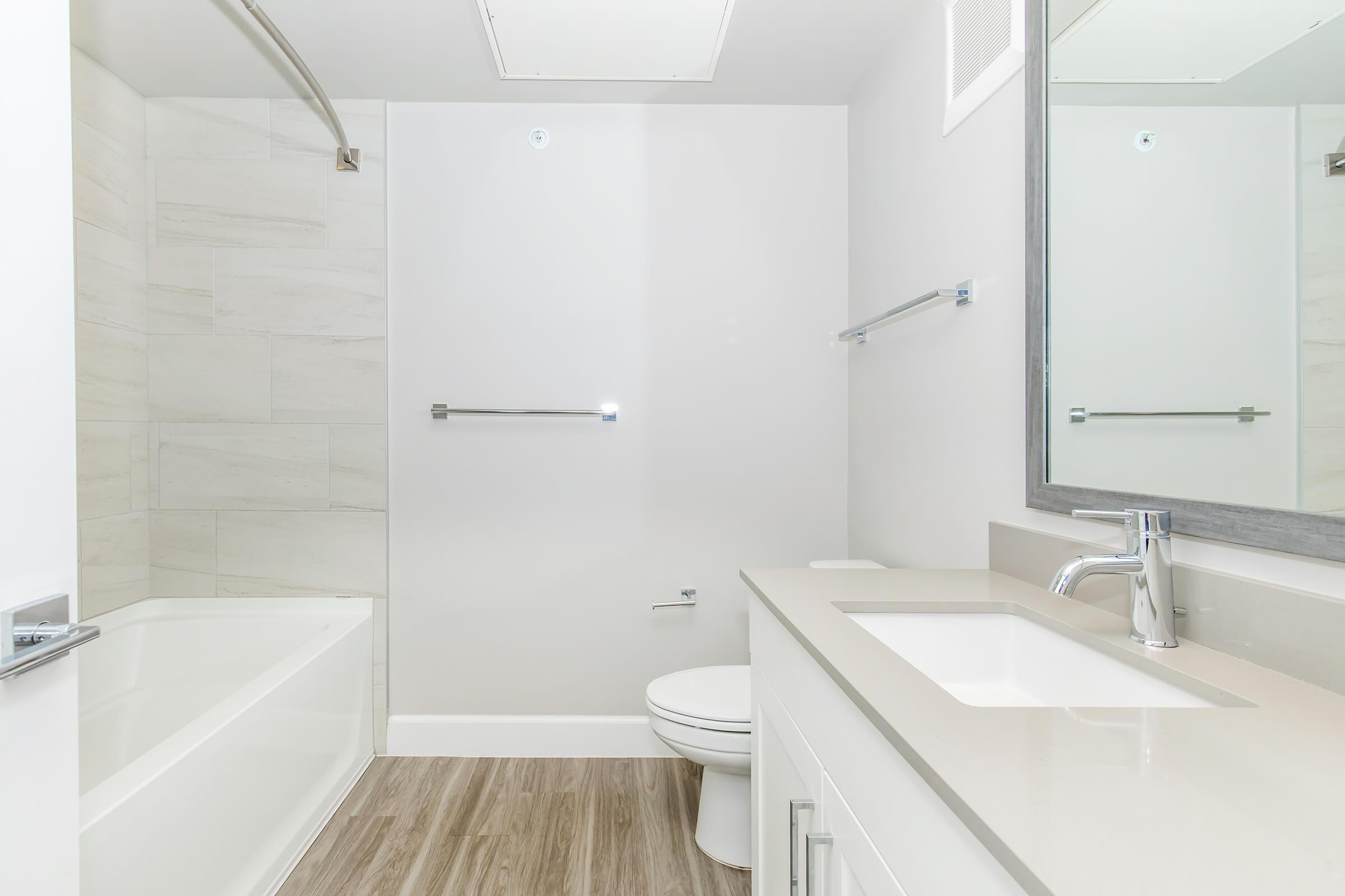 A modern bathroom featuring a white bathtub, a shower with glass partition, a white toilet, and a sleek vanity with a sink. The walls have light-colored tiles, and the floor is a wood-like finish. A large mirror reflects the space, enhancing the openness and brightness of the room.