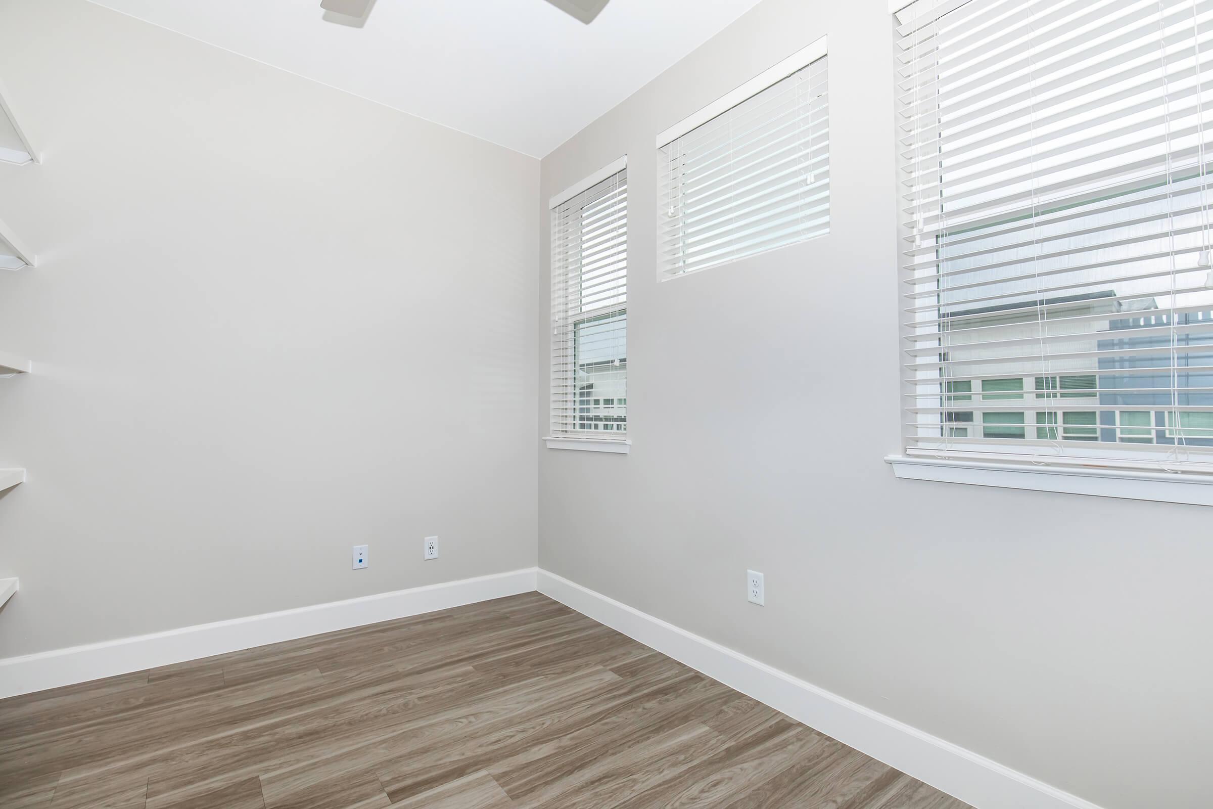 A bright, empty room featuring light gray walls and multiple windows with blinds. The floor is made of wood-like tiles, and there is a ceiling fan overhead. Two walls are devoid of furnishings, highlighting the spaciousness of the area.