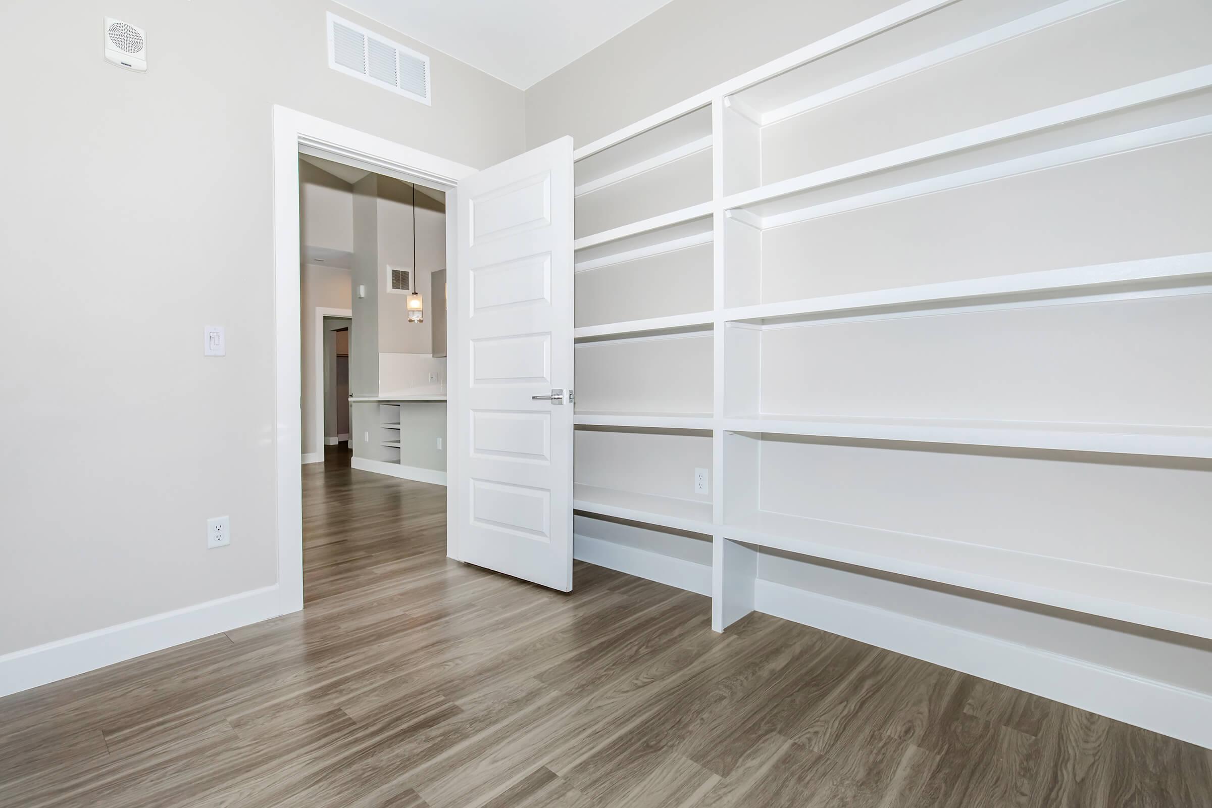 A modern room with light-colored walls and a wooden floor. An open white door leads to another space, while one wall features white built-in shelves. The room is well-lit and has a minimalist design, creating a spacious and inviting atmosphere.