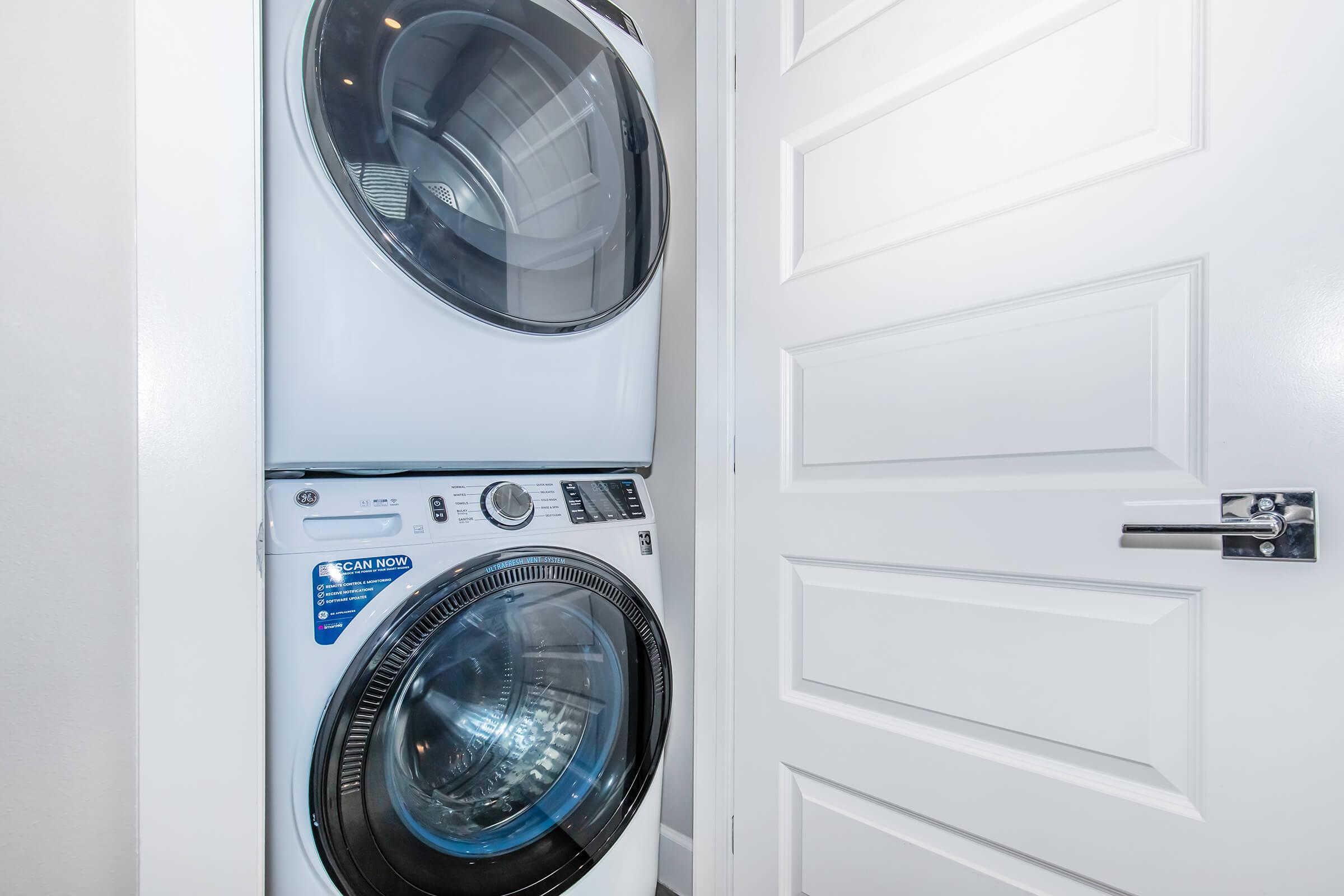 A stacked washer and dryer located in a narrow space, with a white door partially visible in the background. The appliances are modern and sleek, featuring circular doors and control panels. The overall setting is clean and bright, with neutral-colored walls.